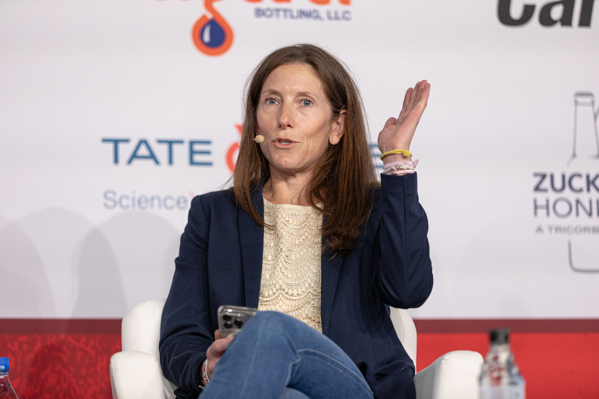 Woman in a navy blazer raises one hand while seated in front of a Tate and Lyle sponsor wall.