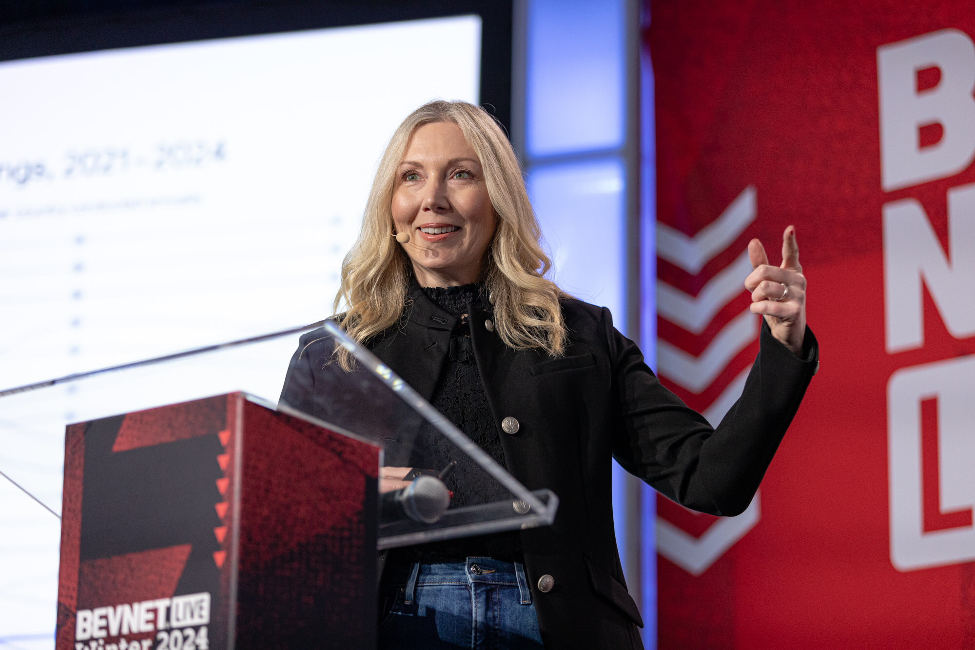 Woman with blond hair in a black jacket gestures at a podium in front of a chart slide.