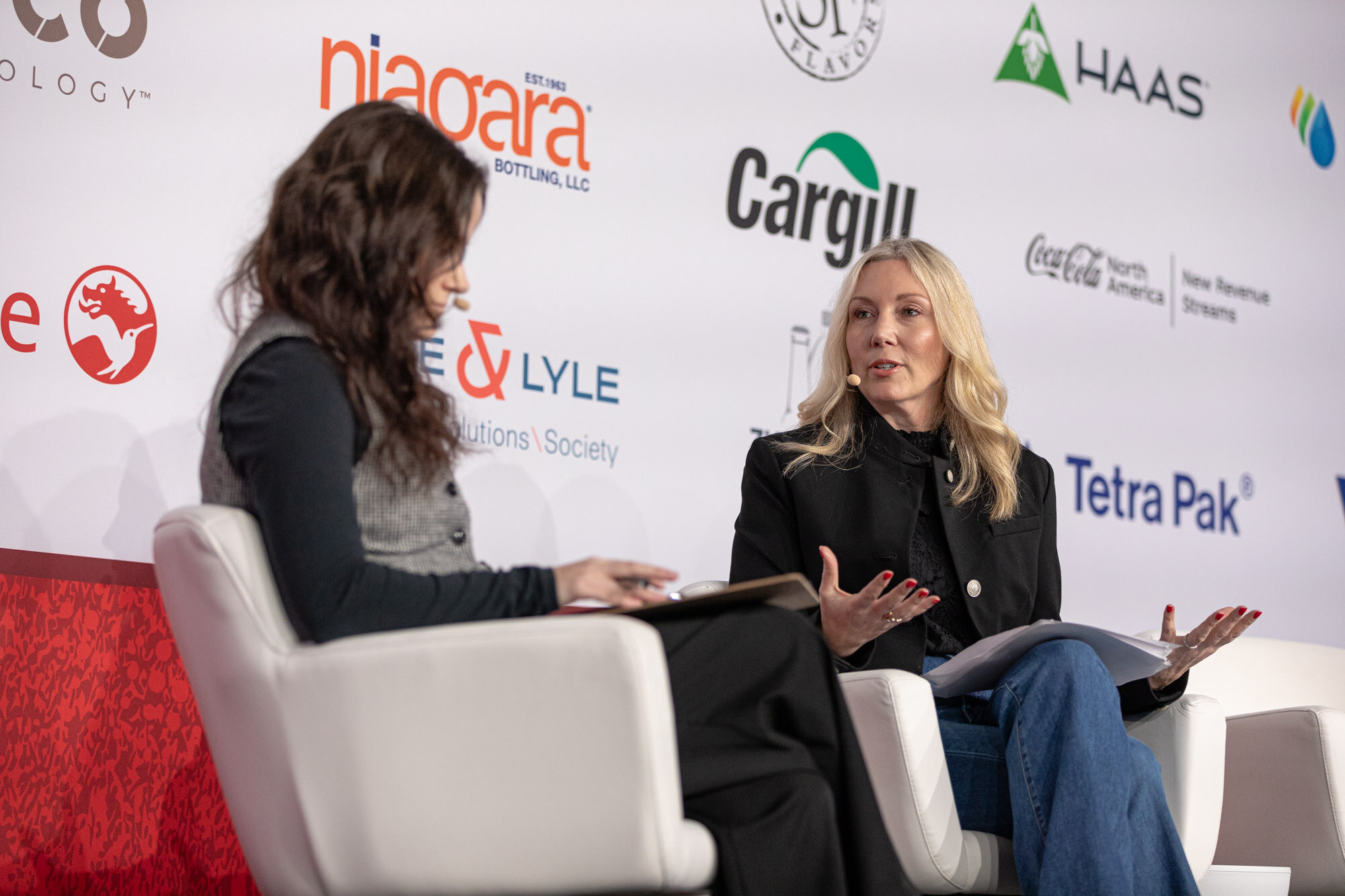 Two women sit on stage in white chairs, one with dark hair and one with blond hair, in front of a sponsor wall.