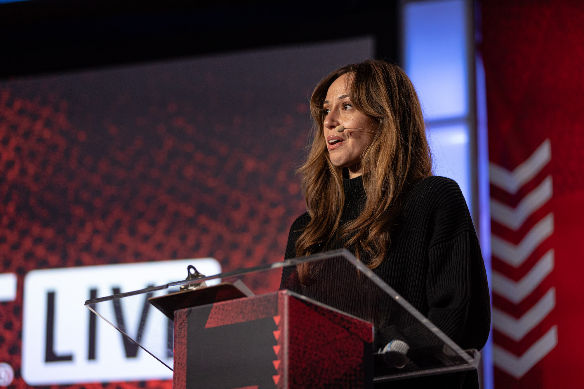 Woman with long hair in black speaks at a podium in front of a red BevNET Live backdrop.