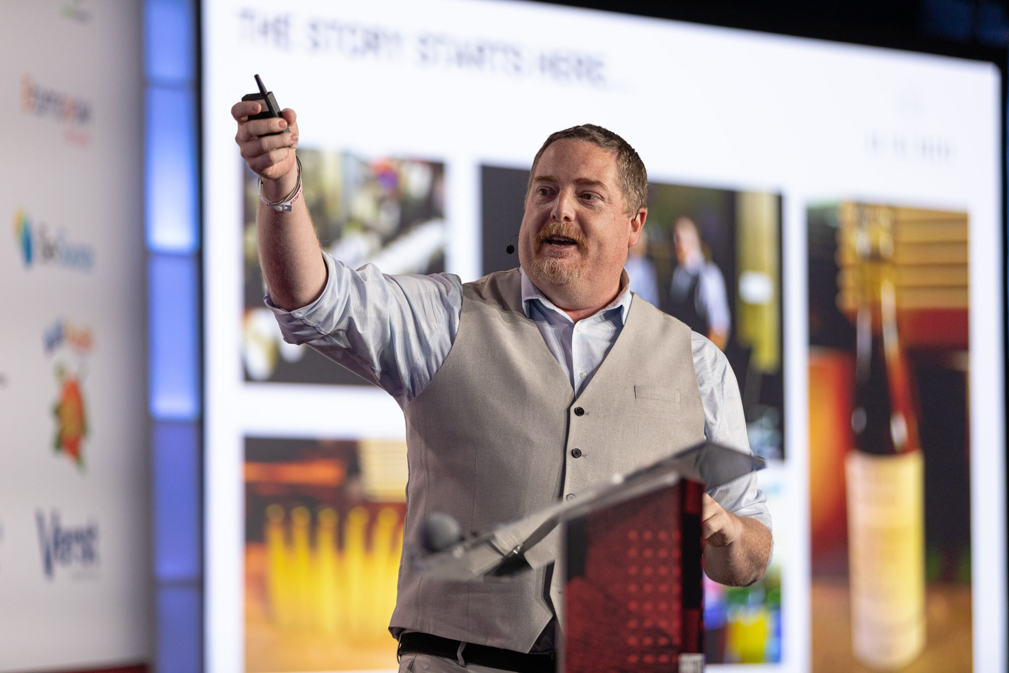 Man in a gray vest holds a clicker up while standing at a lectern in front of a slide of bottles.