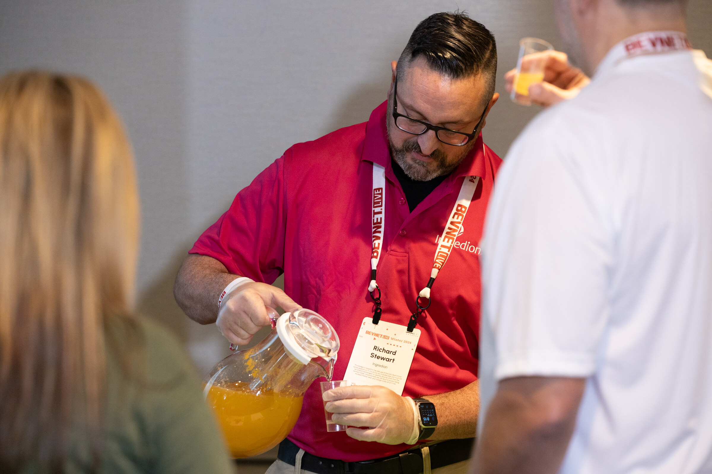 Man in a red shirt and glasses pours juice from a pitcher into a cup while others stand around him.