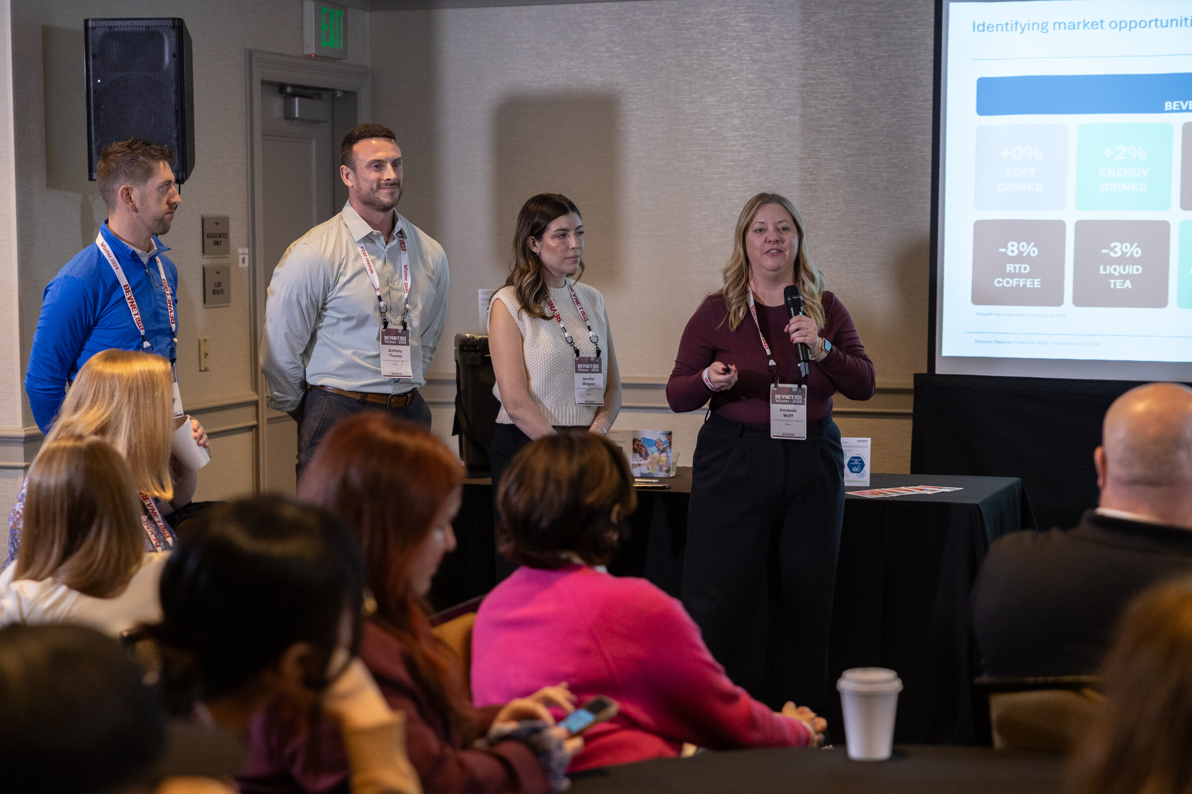 Four people stand at the front of a meeting room next to a screen while attendees sit in the foreground.
