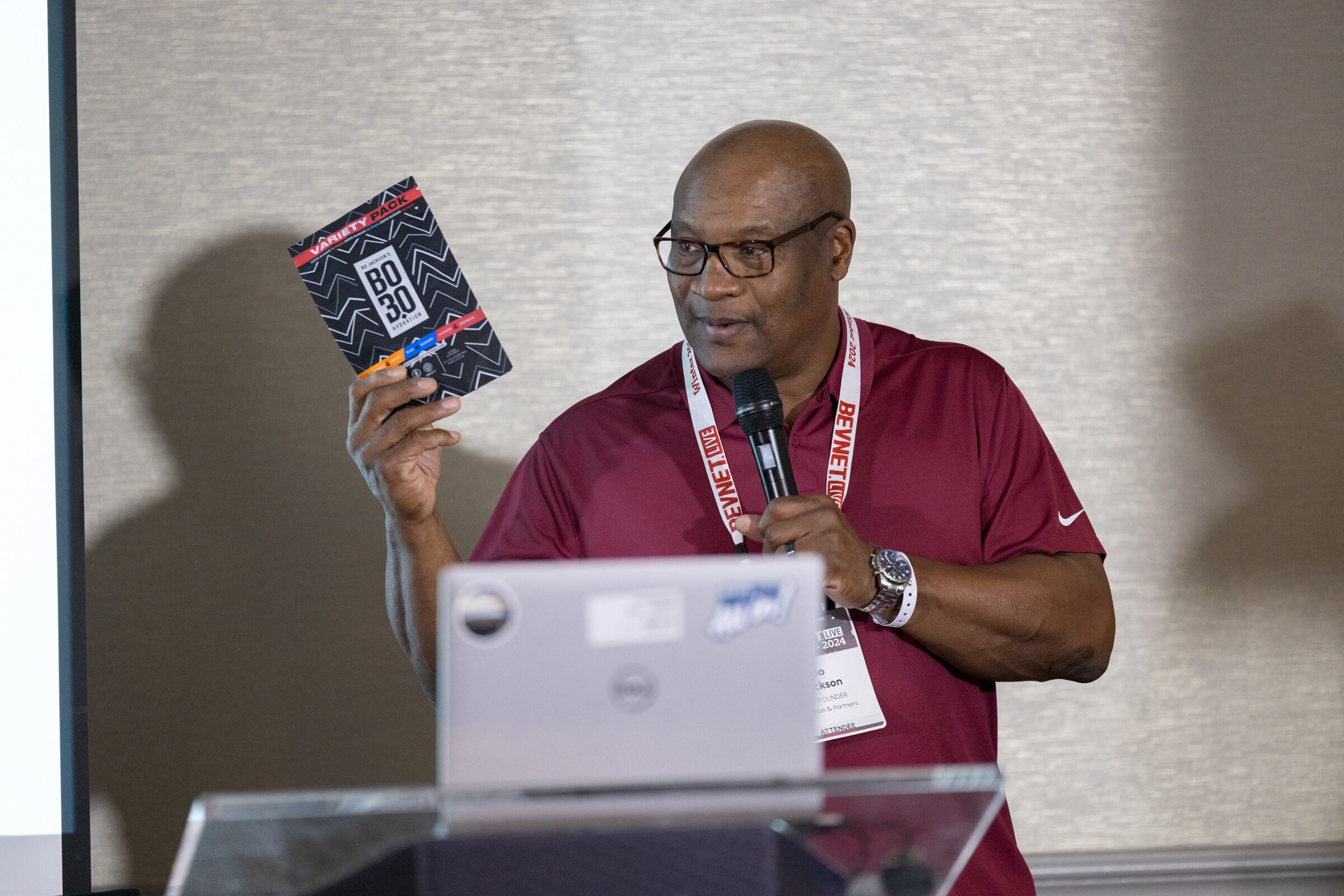 Man in a maroon polo holds up a card and a microphone beside a laptop on a podium.