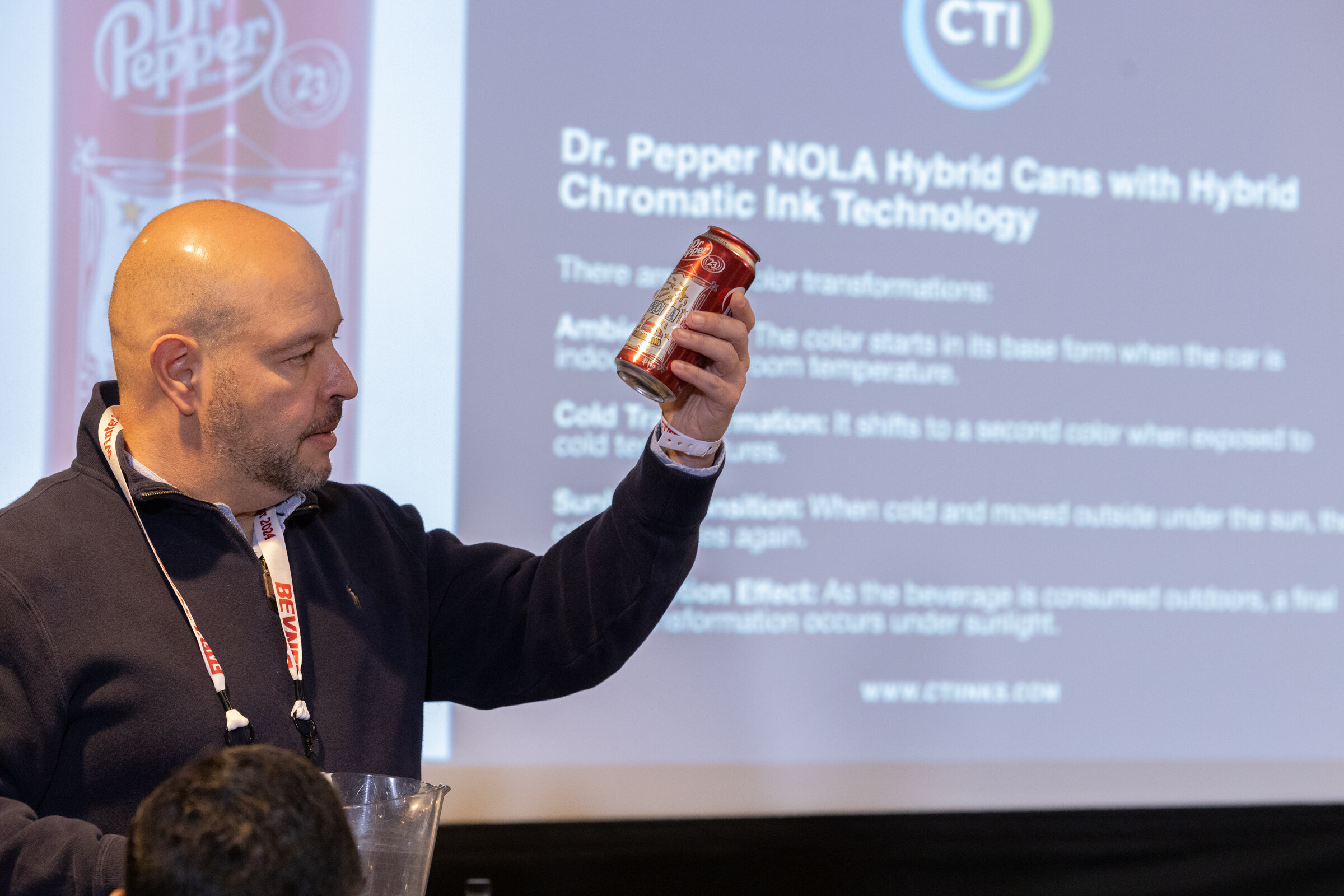 Bald man in a navy pullover holds up a red can in front of a slide that reads Dr. Pepper NOLA Hybrid Cans with Hybrid Chromatic Ink Technology.