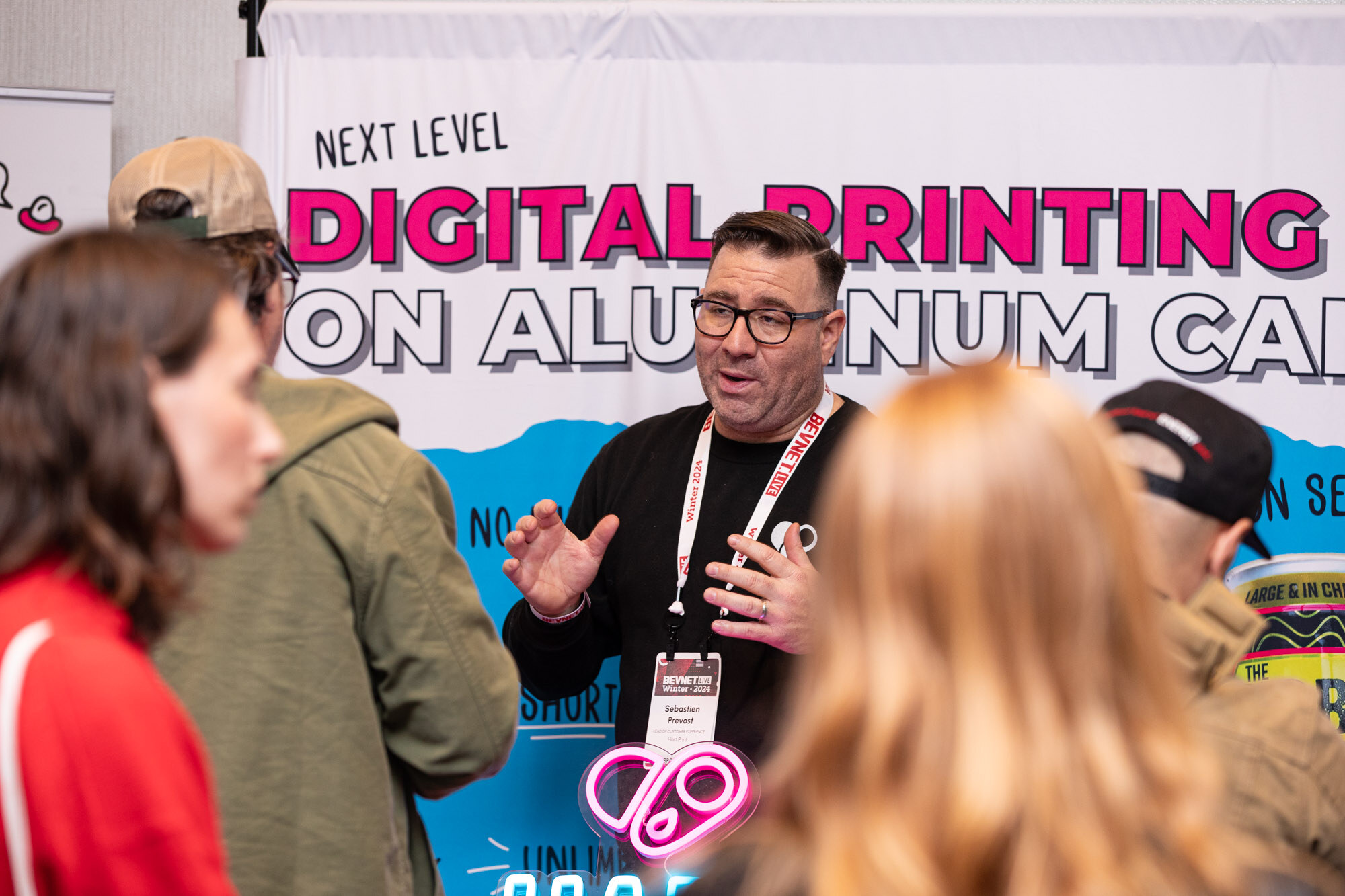 Man in a black shirt and glasses gestures in front of a backdrop reading Digital Printing on Aluminum Cans.