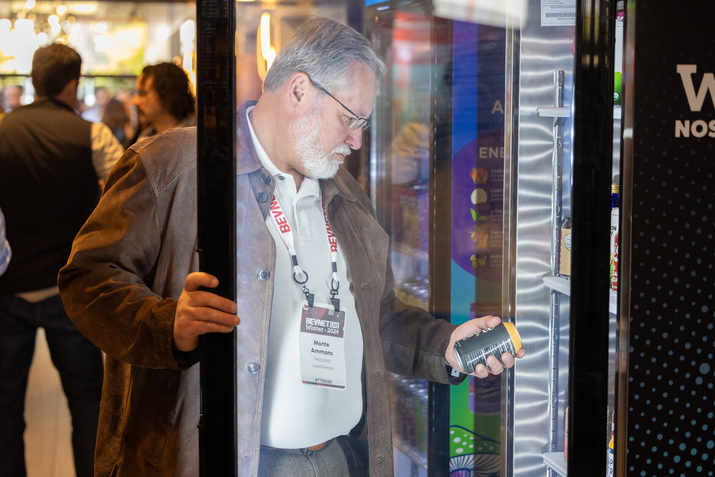 Man with glasses and a lanyard opens a glass cooler door and holds a can.