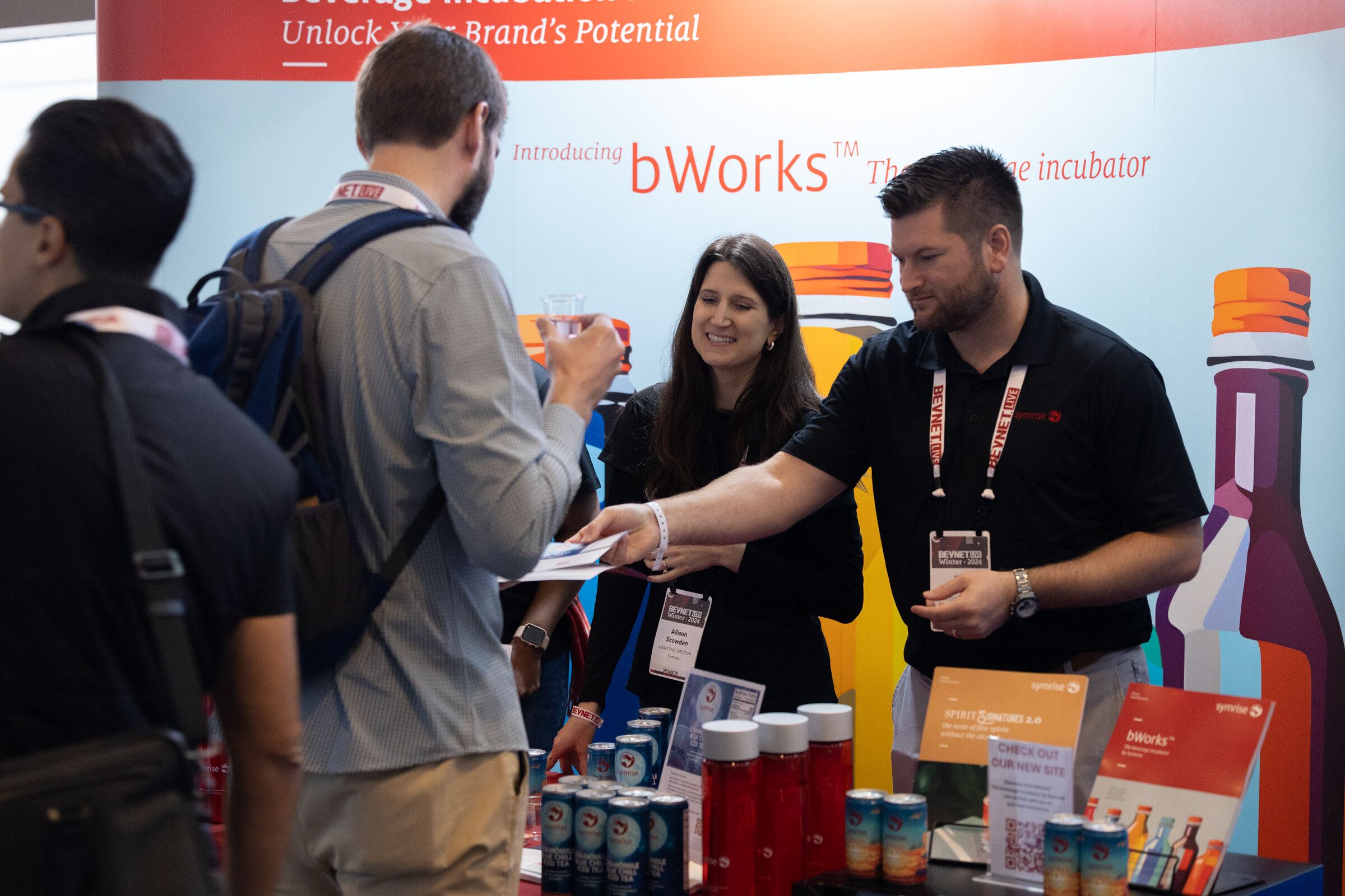 Several attendees talk in front of a bWorks display with colorful bottles, one man hands over a card.