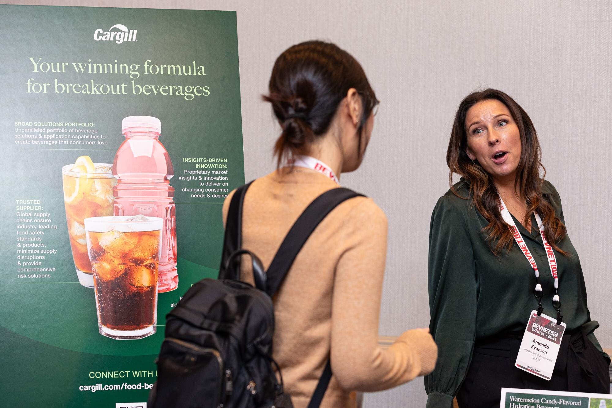 Two women with lanyards talk in front of a green Cargill poster showing a pink drink and an iced cola.