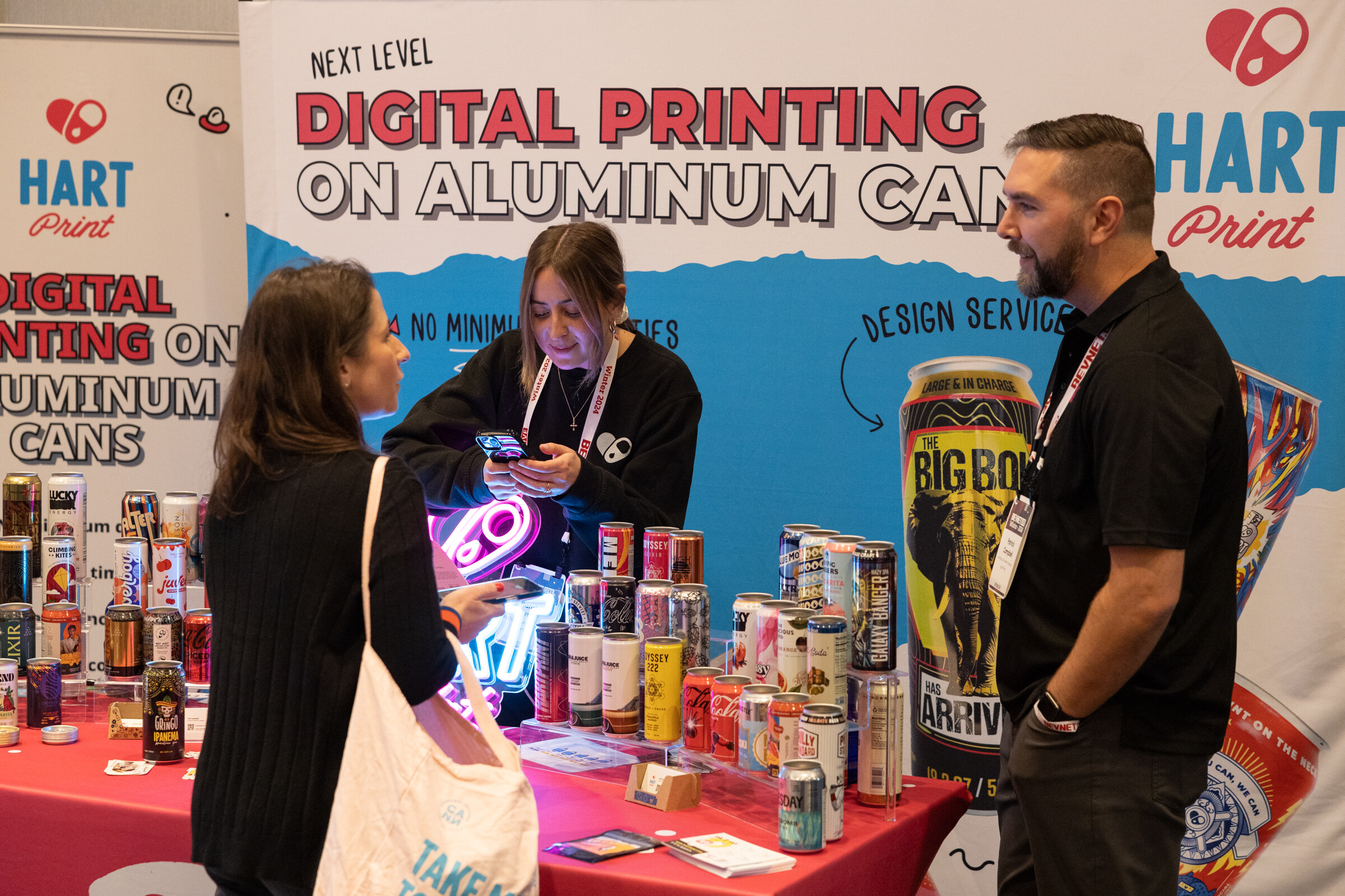 A woman and two men stand at a pink table covered in printed cans in front of a Digital Printing on Aluminum Cans backdrop.