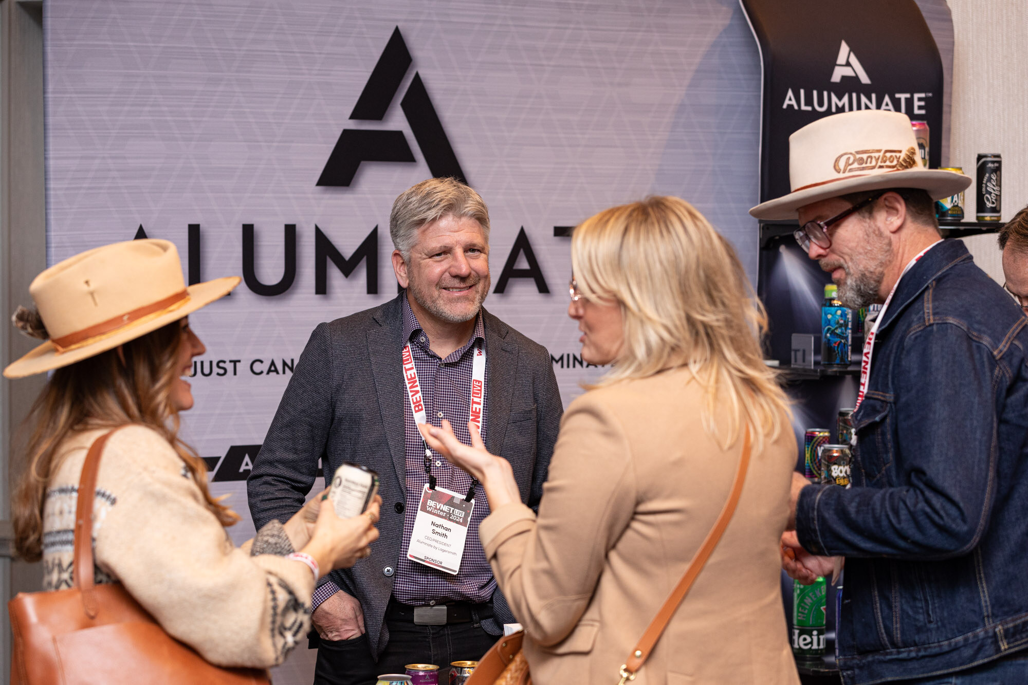Two men and two women, one in a cowboy hat, talk in front of a black Aluminate backdrop.