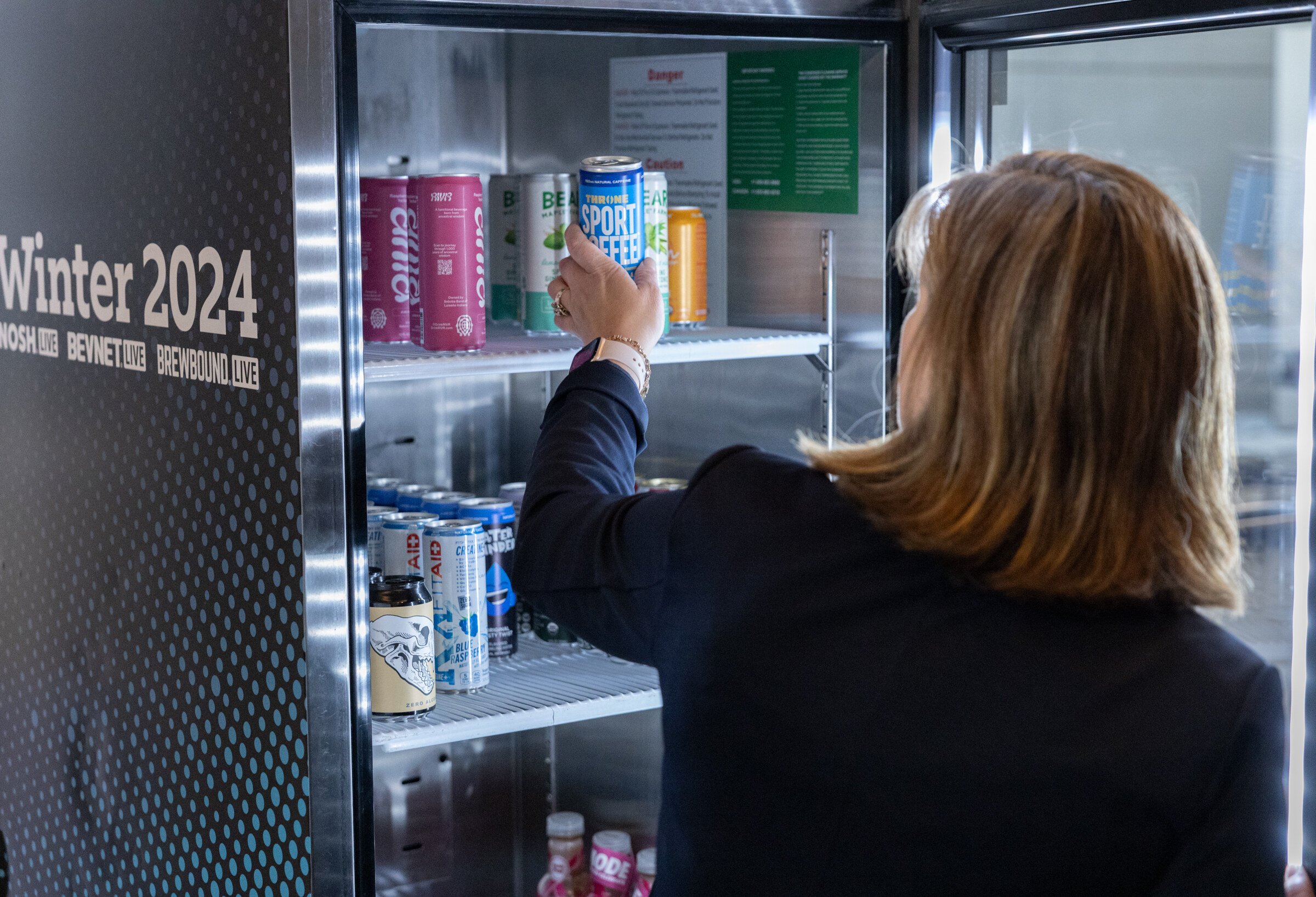 Woman reaches into a glass-door fridge stocked with cans and bottles under a Winter 2024 sign.