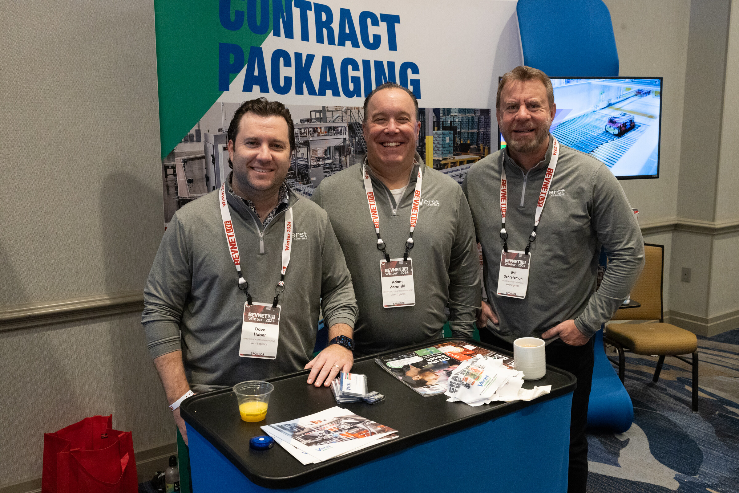 Three men with lanyards smile behind a blue table covered in brochures.