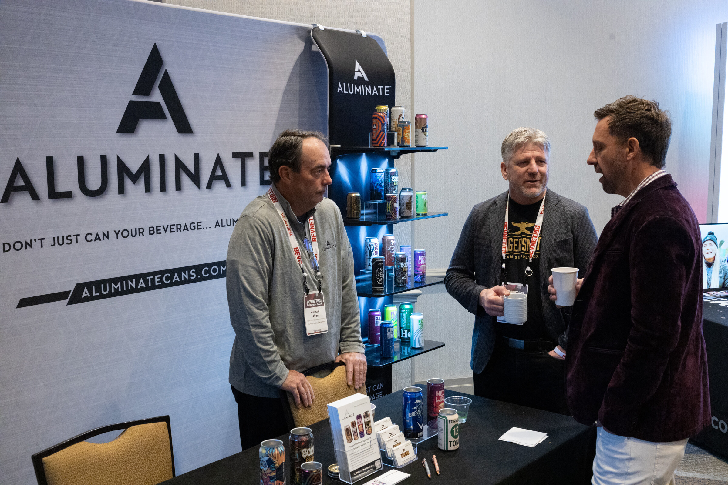 Three men stand in front of a black Aluminate display with cans.