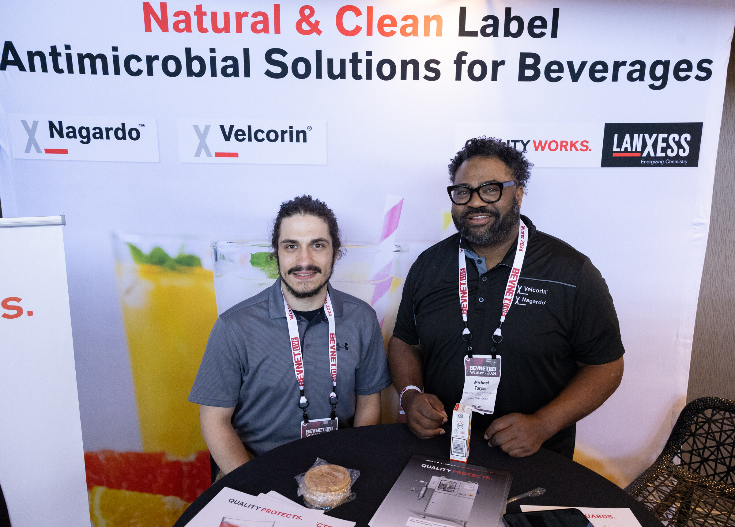 Two men with lanyards smile at a table in front of a banner reading Natural and Clean Label Antimicrobial Solutions for Beverages.