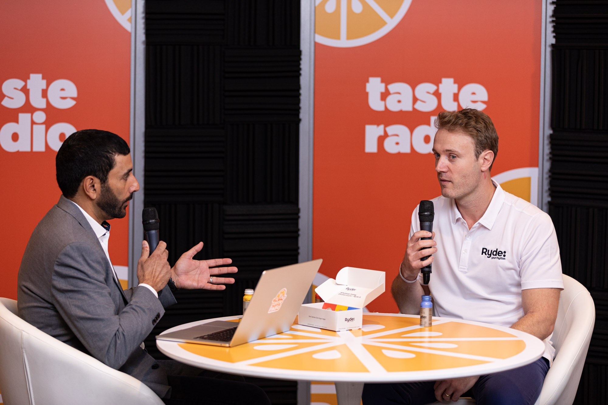 A man in a gray blazer and a man in a white Ryde polo sit at an orange round table in front of a Taste Radio backdrop.