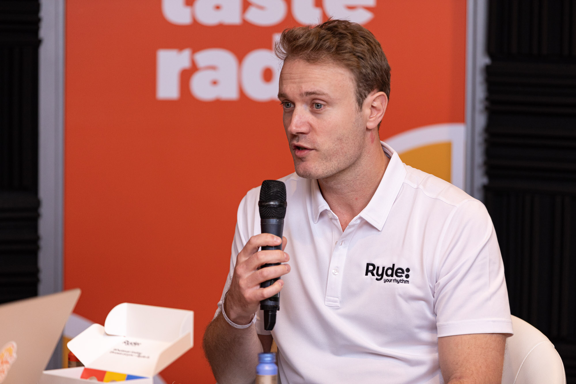 Man in a white Ryde polo holds a microphone in front of an orange Taste Radio banner.