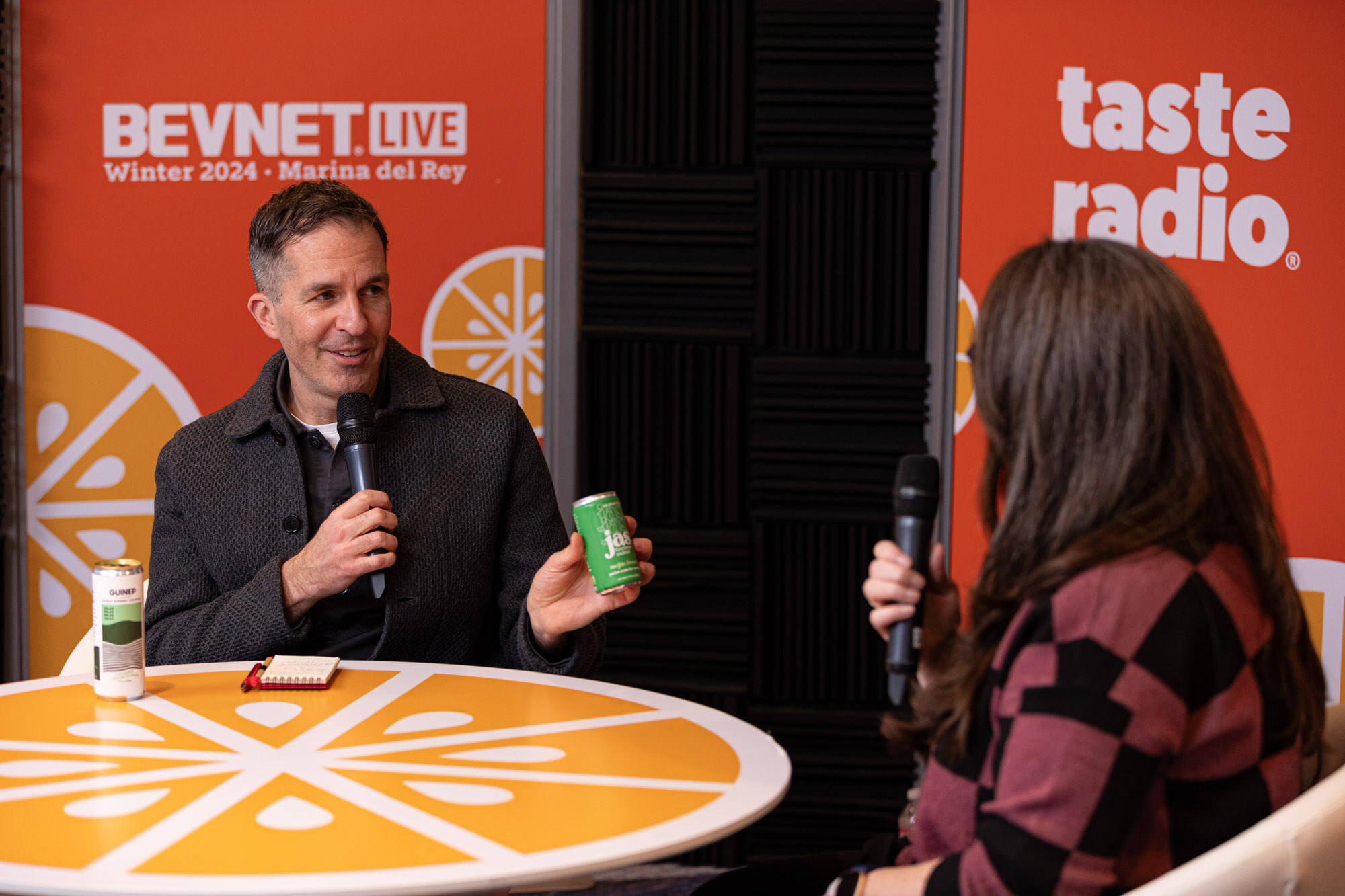 A man in a dark jacket holds a green can at a round orange table beside a BevNET Live Winter 2024 banner.