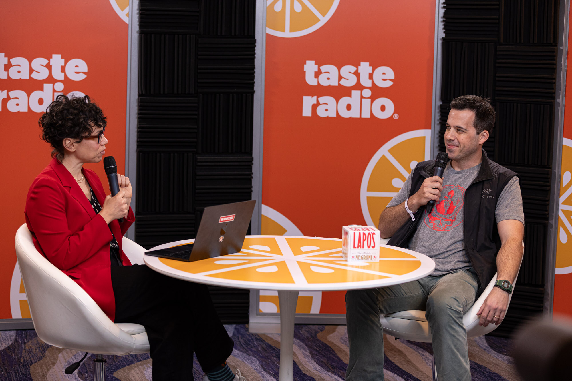 A woman in a red blazer and a man in a gray t-shirt and black vest sit at an orange round table with a laptop and a Lapos box.