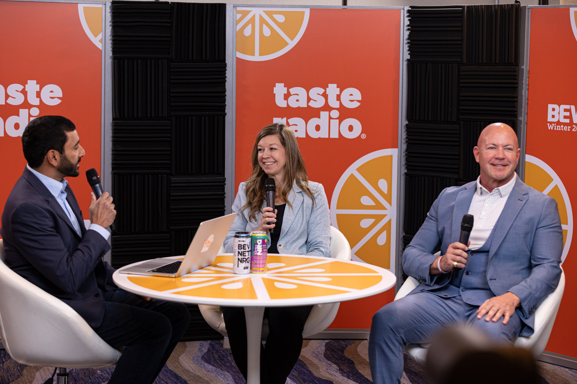 A man in a navy suit, a woman in a light blue blazer and a man in a blue suit sit at an orange round table with microphones.
