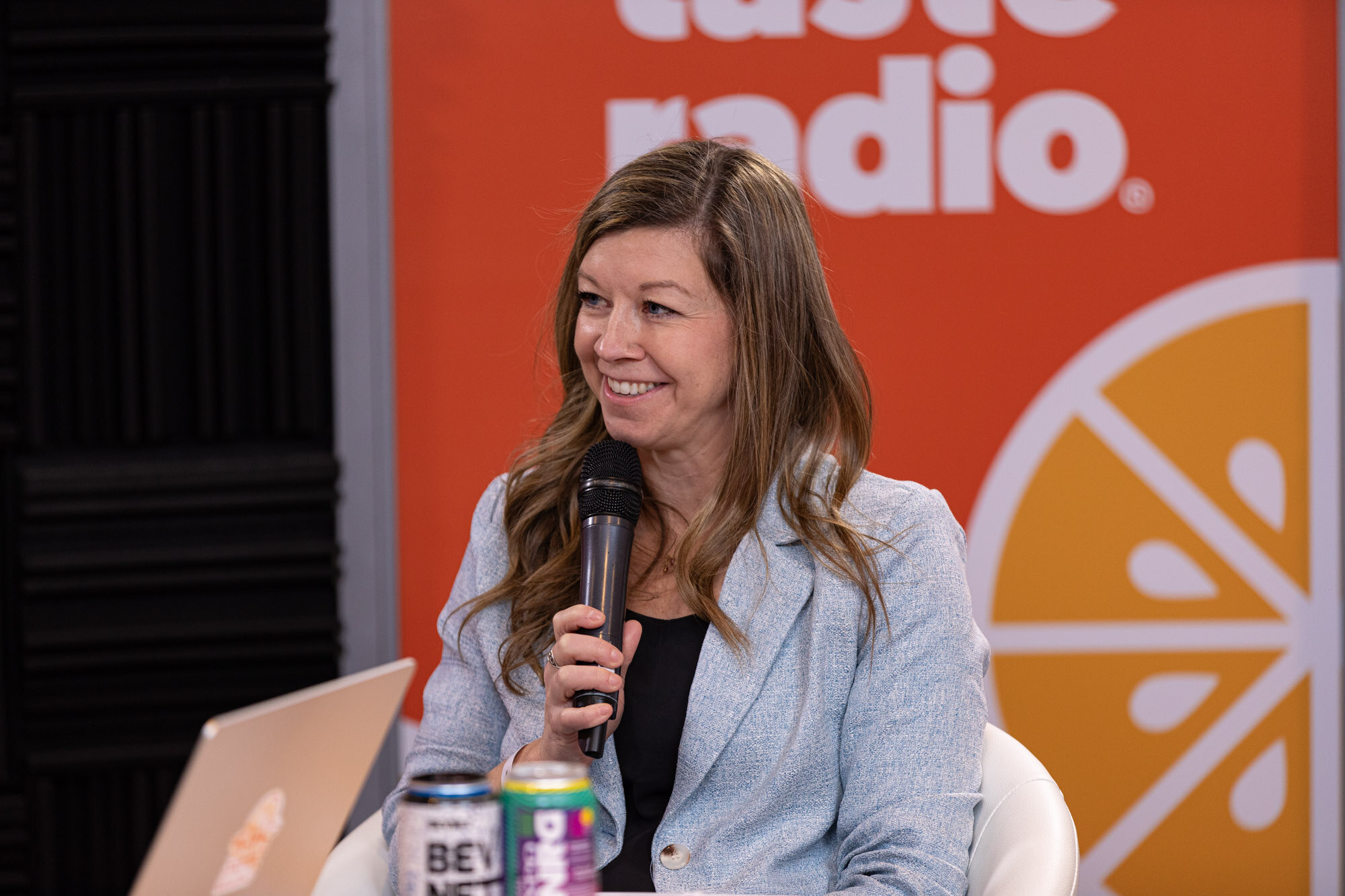 Smiling woman in a light blue blazer holds a microphone in front of an orange Taste Radio banner with cans on the table.