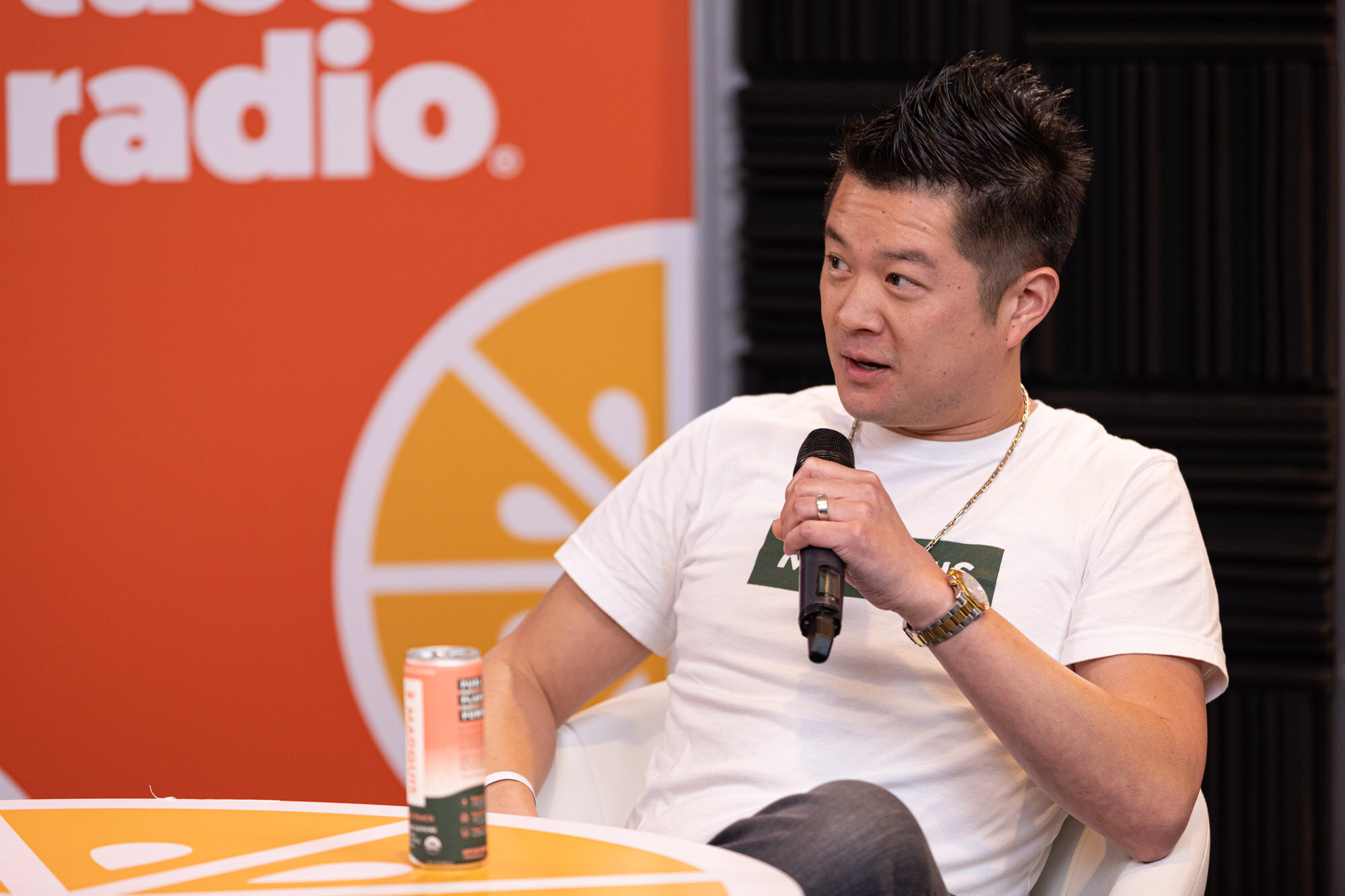 Man in a white t-shirt holds a microphone beside an orange can on an orange Taste Radio table.