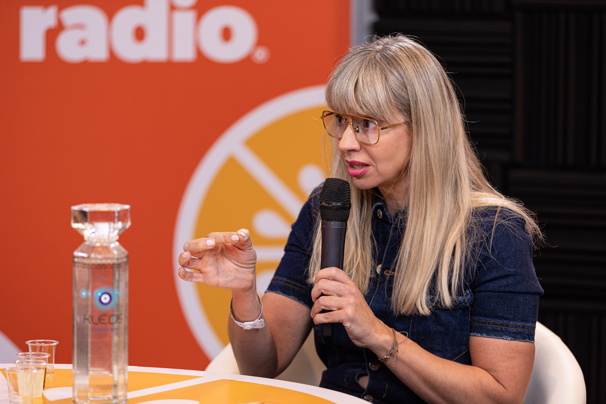 Woman with blond bangs in a denim dress holds a microphone with a glass water bottle on the table in front of her.