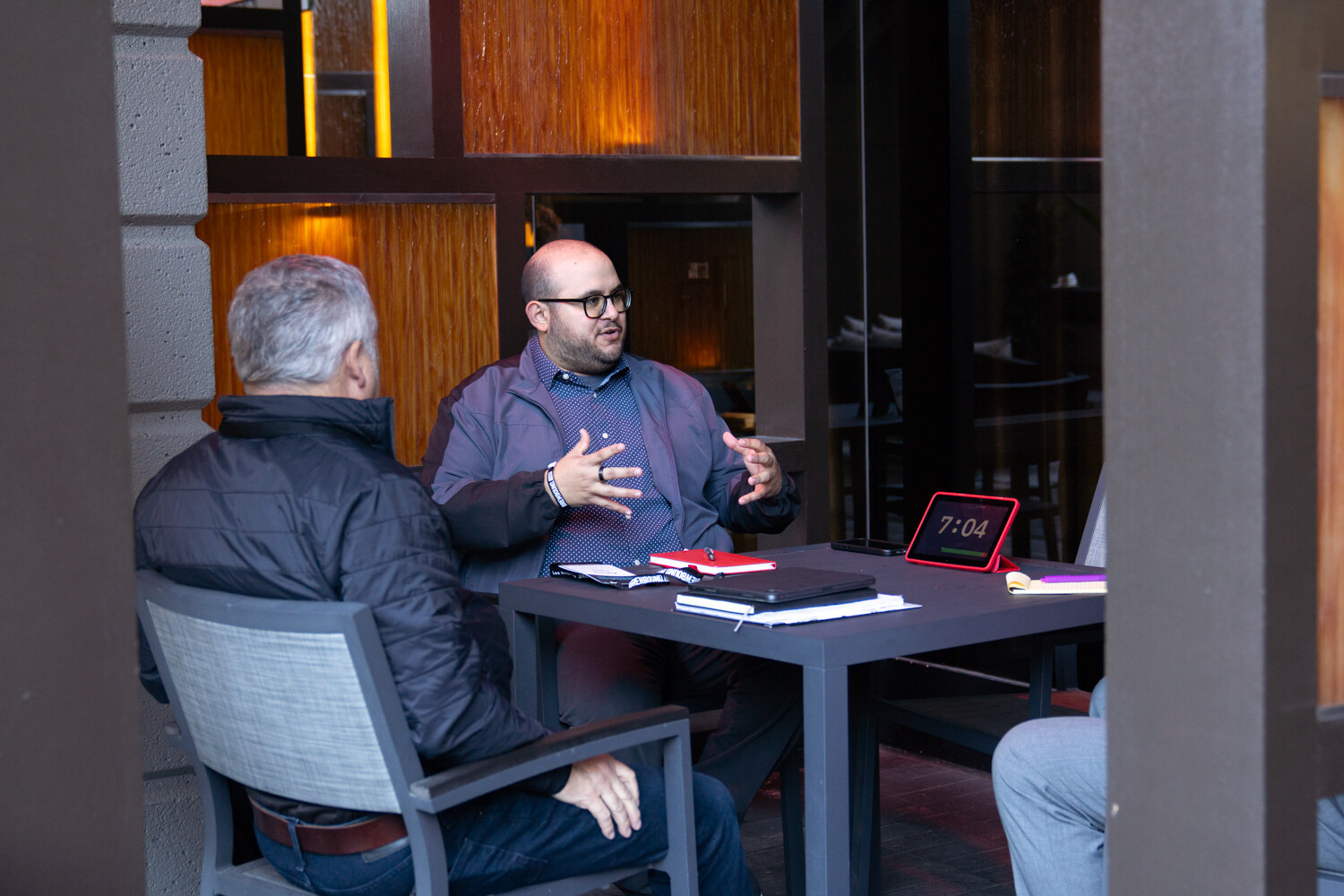 Two men talking across a small table on a patio, one gesturing, with a timer beside a notebook.