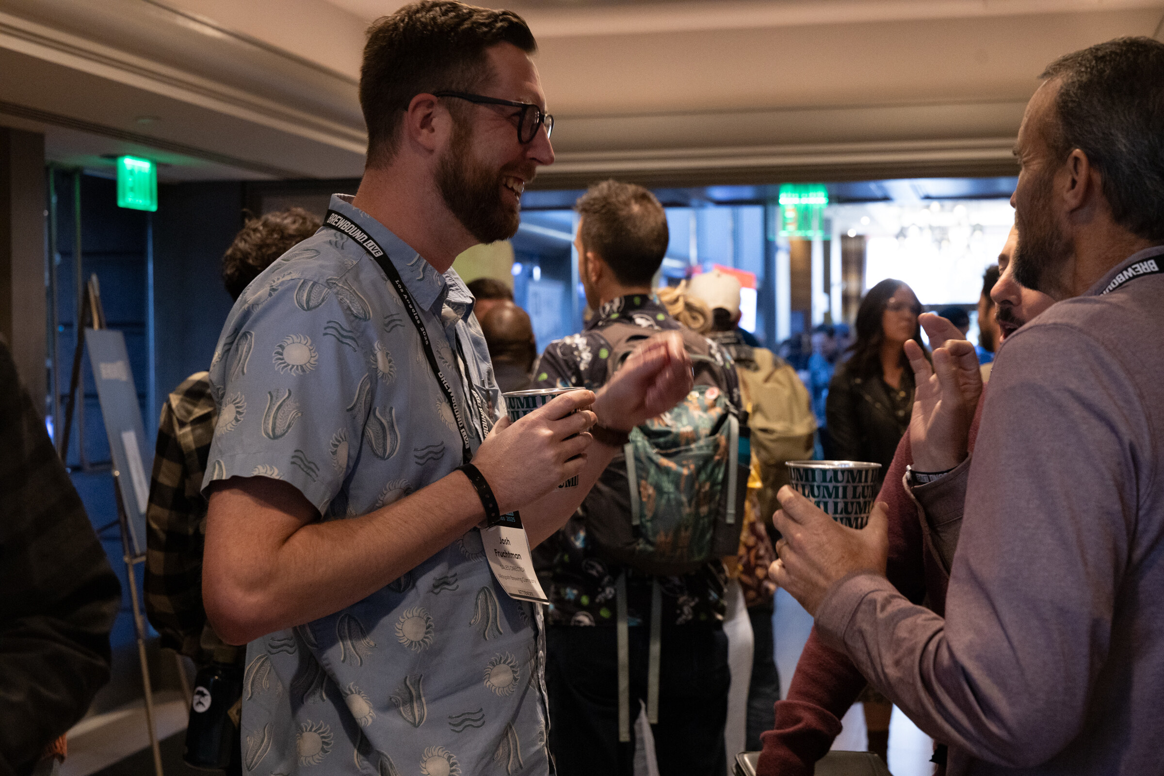Bearded man in a patterned shirt and glasses talking with a man in a mauve sweater, with other attendees behind them.