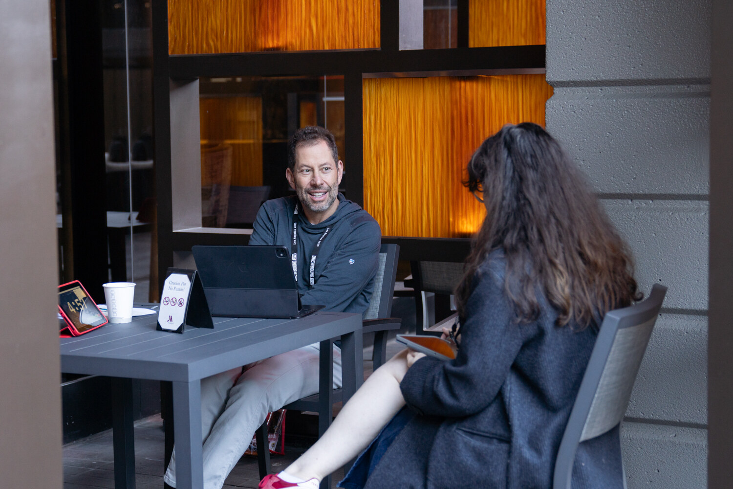 Smiling man in a quarter-zip pullover with a tablet speaking with a woman seen from behind, at an outdoor table.