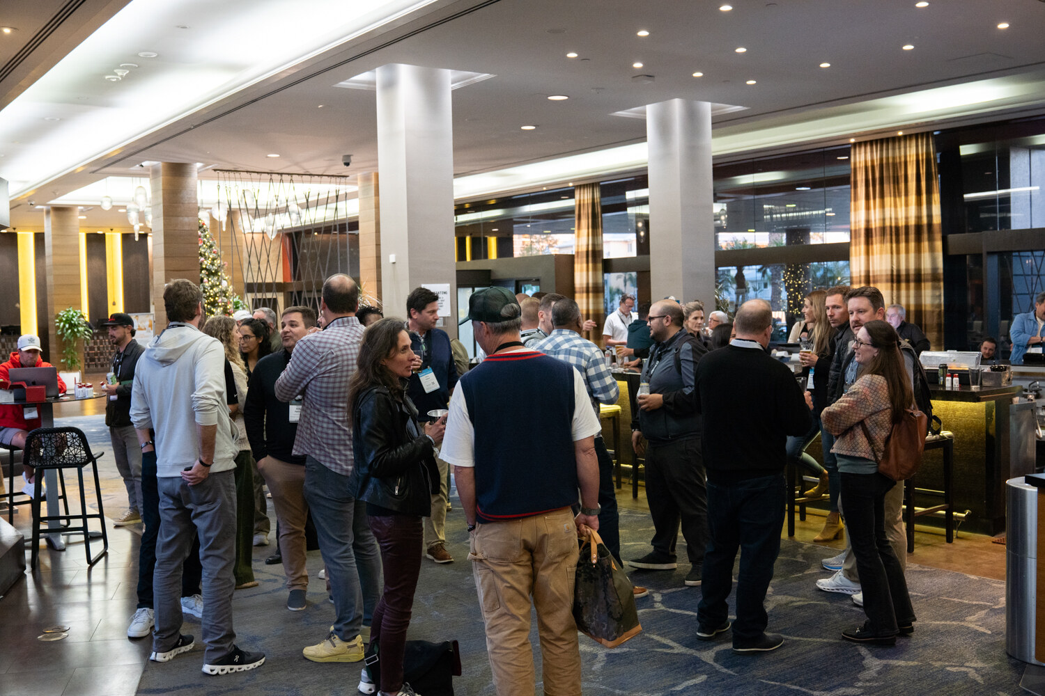 Groups of attendees talking in a hotel lobby with tall columns, a bar and holiday decor.