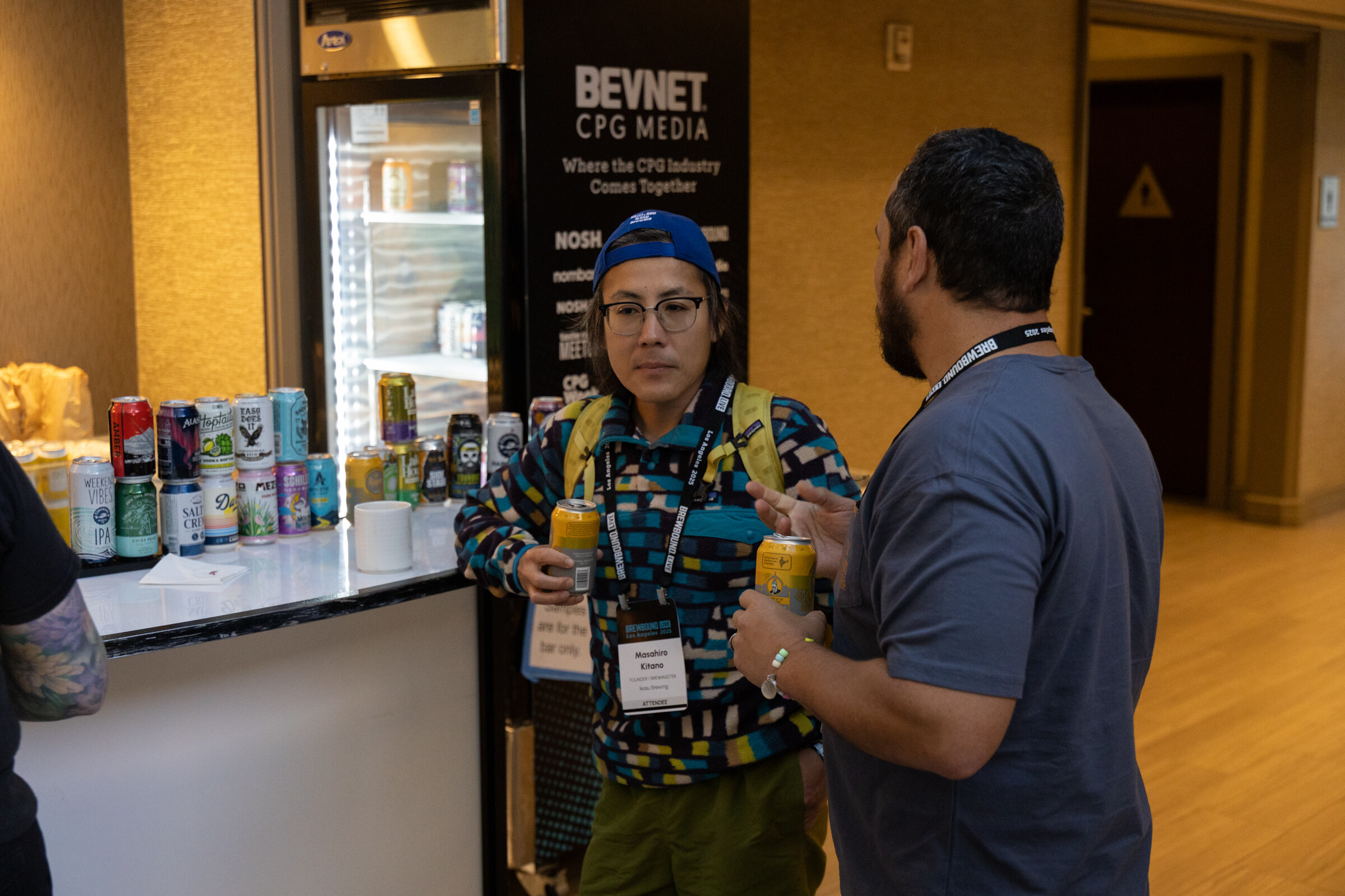 Man in a colorful patterned jacket holding a can and a man in a gray T-shirt, in front of a bar counter lined with cans and a BevNET CPG Media banner.