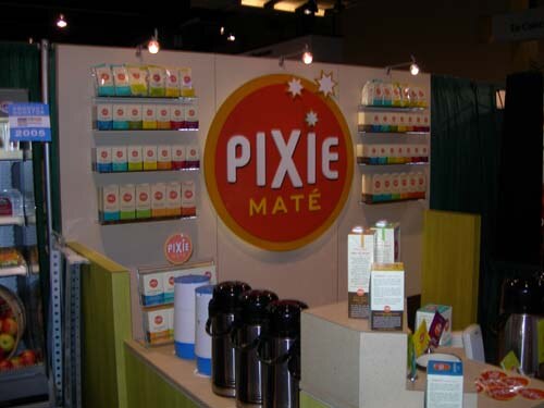 Booth with a large round red Pixie Maté sign, shelves of small boxed products on the walls, and insulated pump pots and product boxes on the counter.