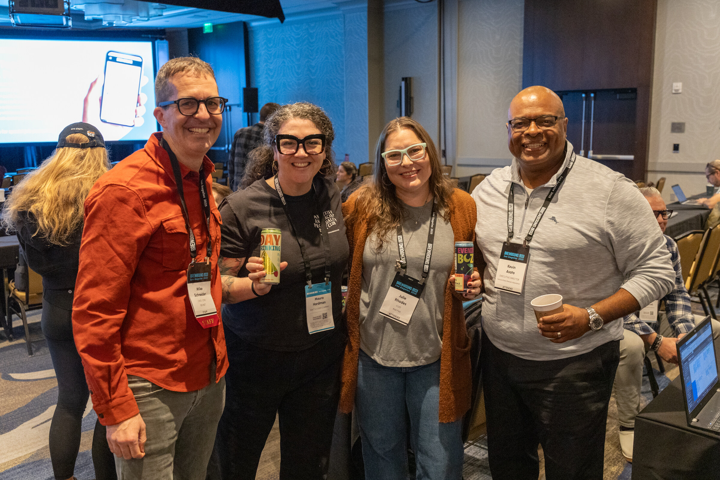 Four smiling attendees with conference lanyards, two holding cans, posing together in a meeting room.