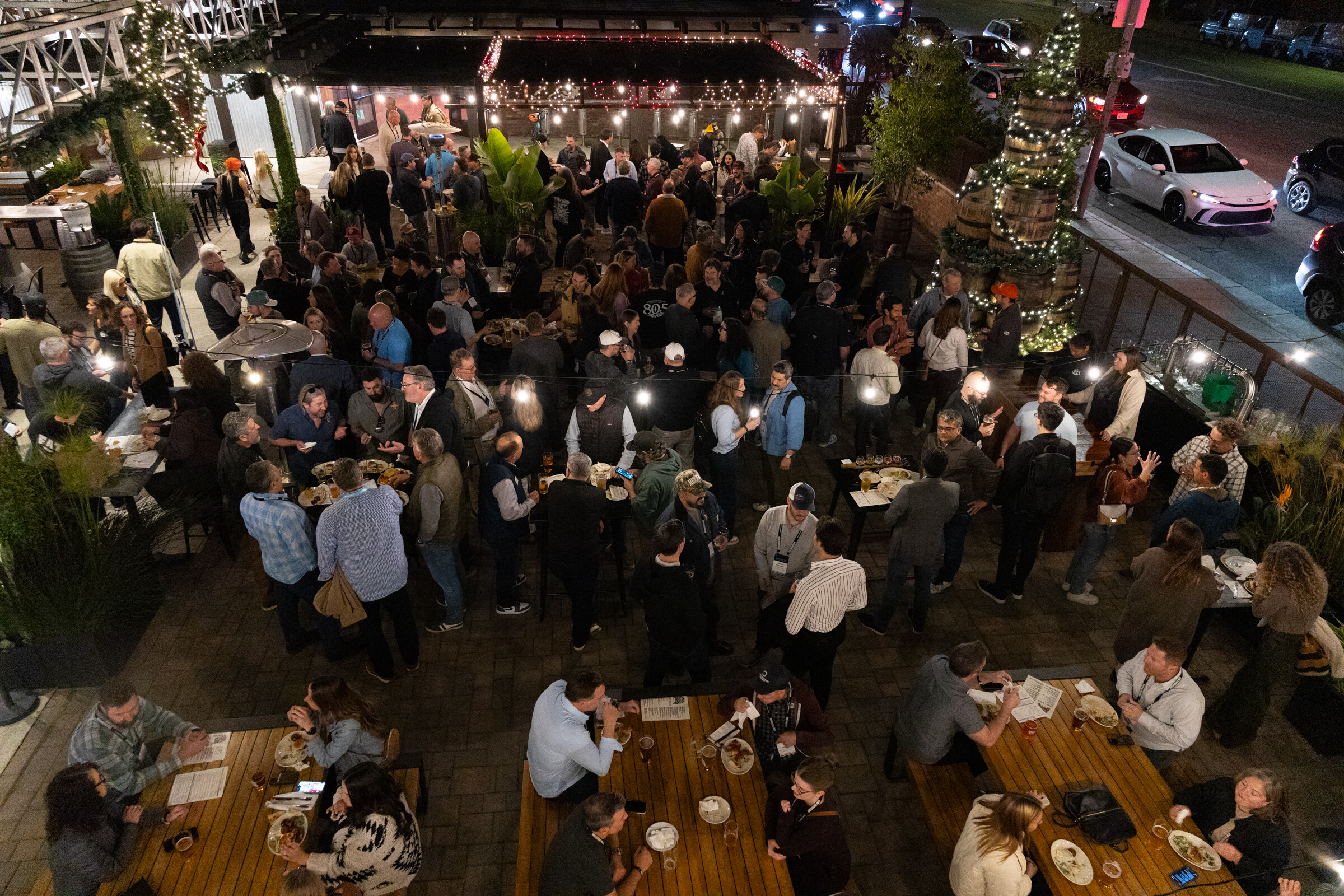 Aerial view of a crowded patio with string lights, wooden tables and guests standing and talking at night.