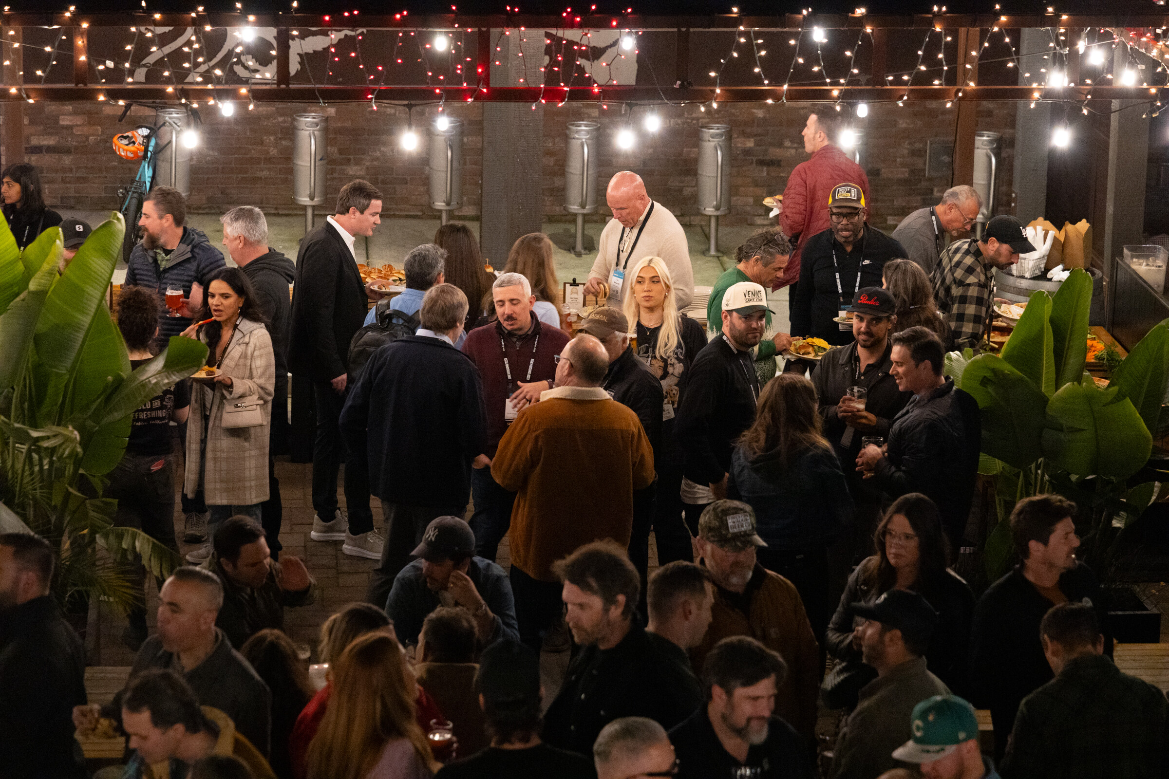 Crowd of attendees standing and talking on a lit patio with plants and string lights overhead.