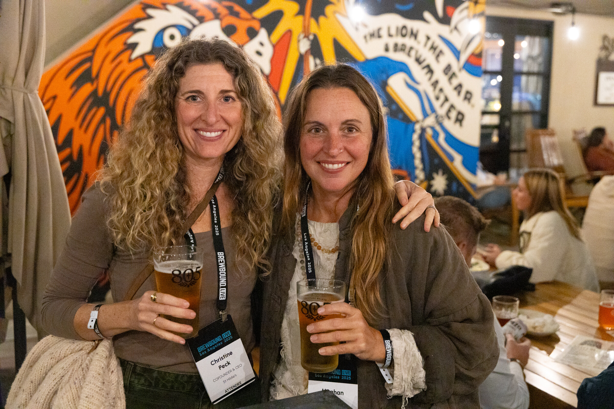 Two smiling women with conference lanyards holding pints of beer in front of a tiger mural.