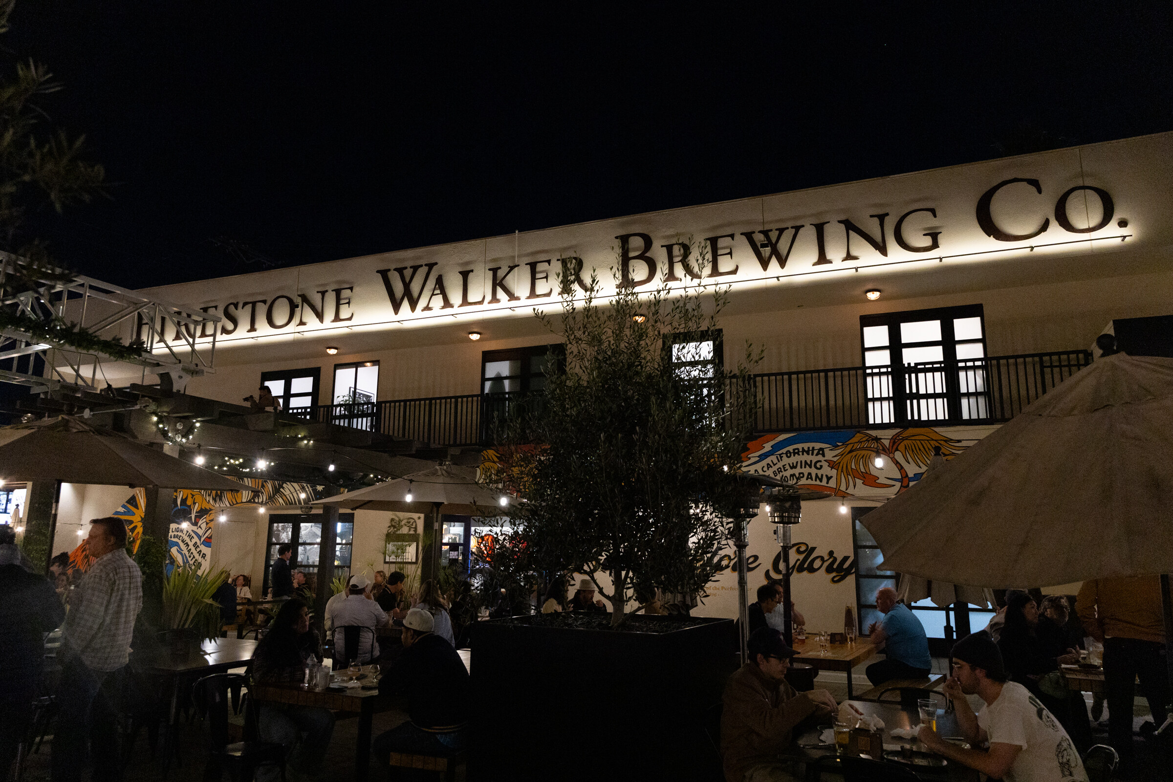 Exterior of a building with a Firestone Walker Brewing Co. sign at night, with guests at patio tables below.