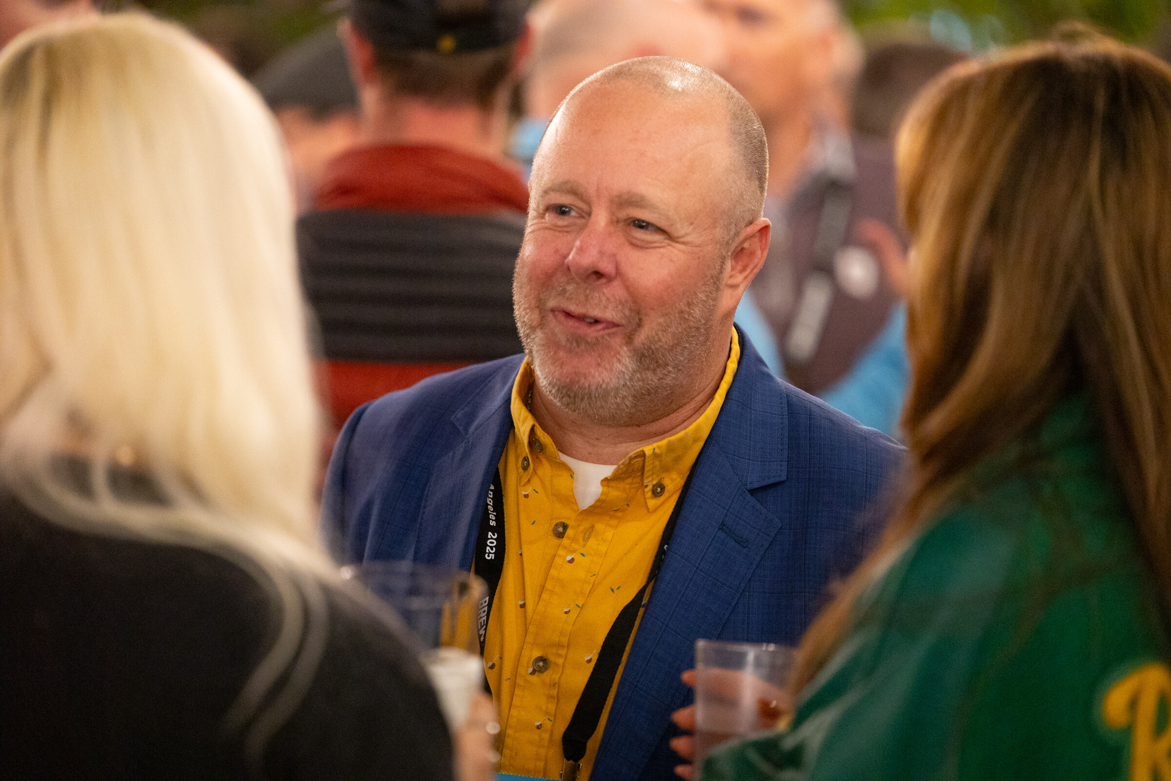 Smiling bald man in a blue blazer and yellow shirt talking with two women in a crowd.