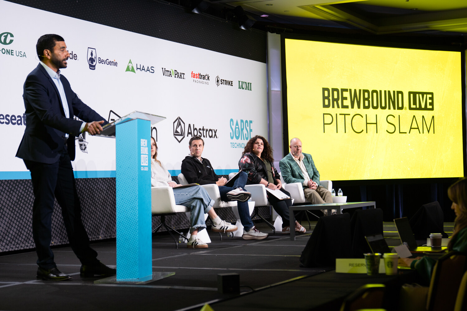 Man in a navy suit at a teal podium on stage with four judges seated on white chairs and a yellow Brewbound Live Pitch Slam screen behind them.