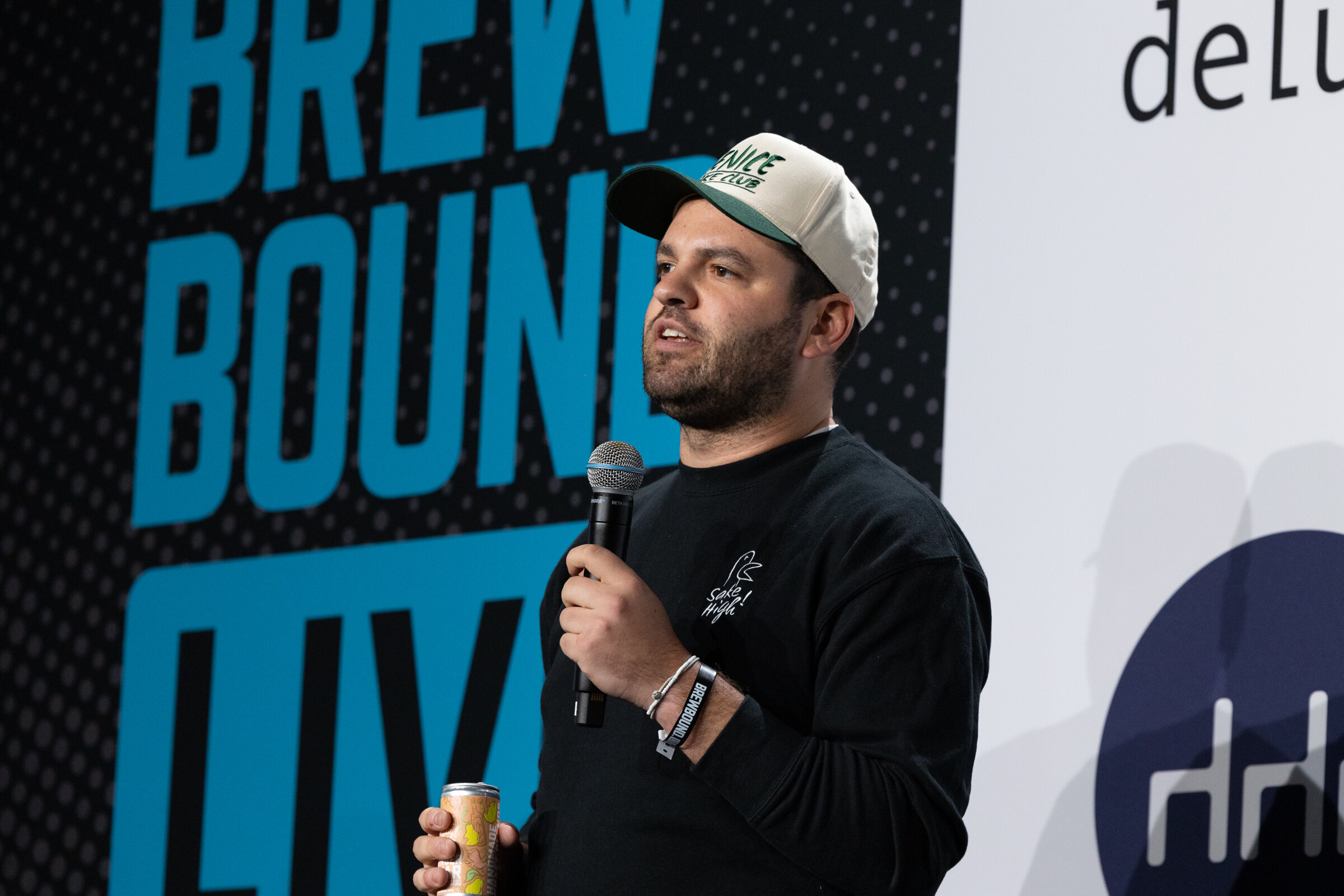 Man in a cream cap and black sweatshirt speaking into a microphone and holding a can on stage in front of a Brewbound Live backdrop.