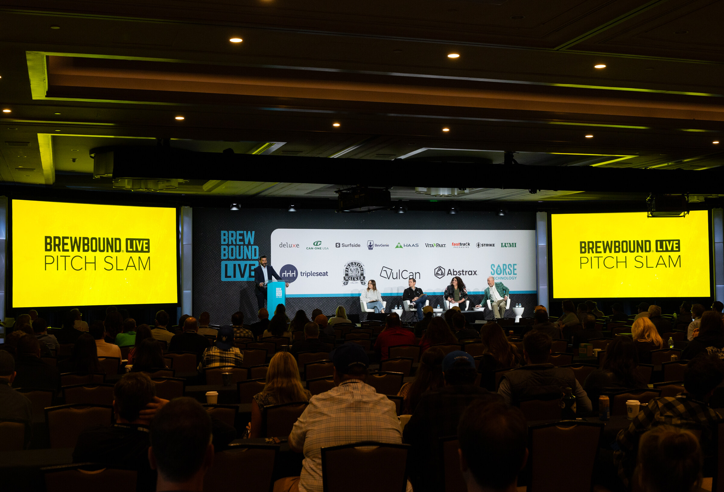 Wide view of a dark ballroom with yellow Pitch Slam screens on either side of the stage and attendees in the foreground.