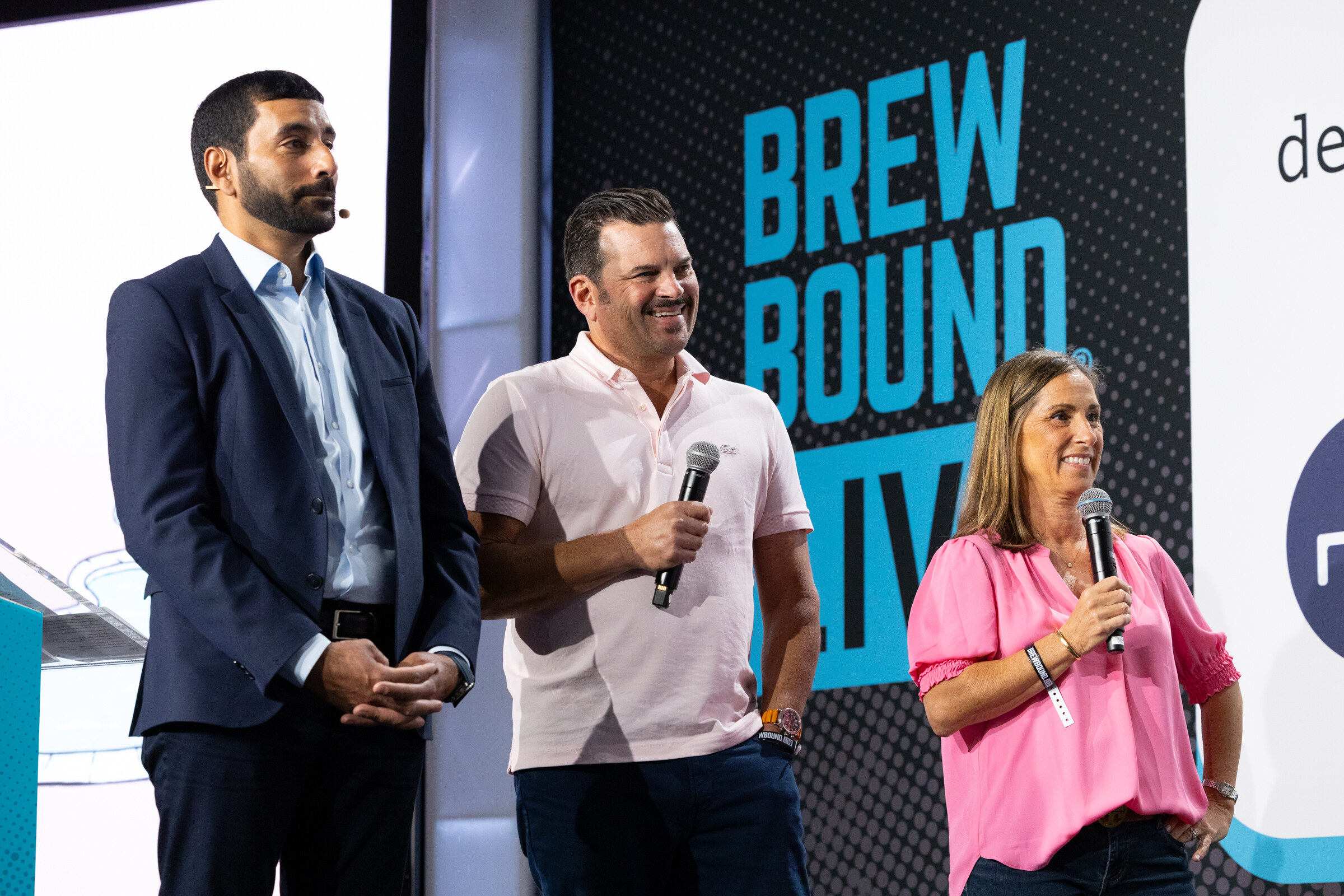 Man in a navy suit, man in a pink polo and woman in a pink blouse holding microphones on stage in front of a Brewbound Live backdrop.