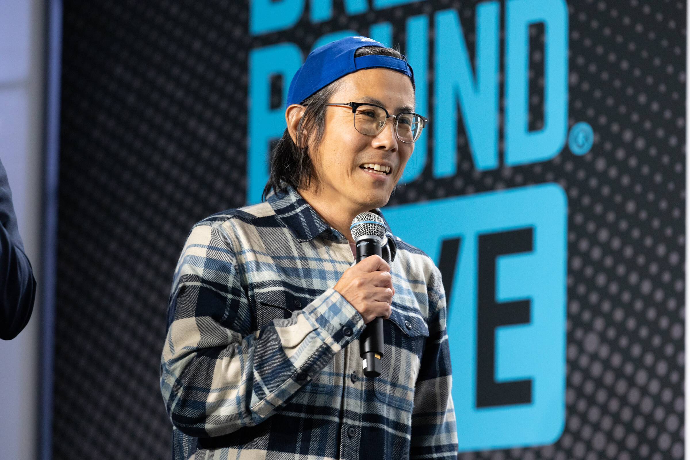 Smiling man in glasses, a blue cap and a plaid flannel shirt speaking into a microphone.