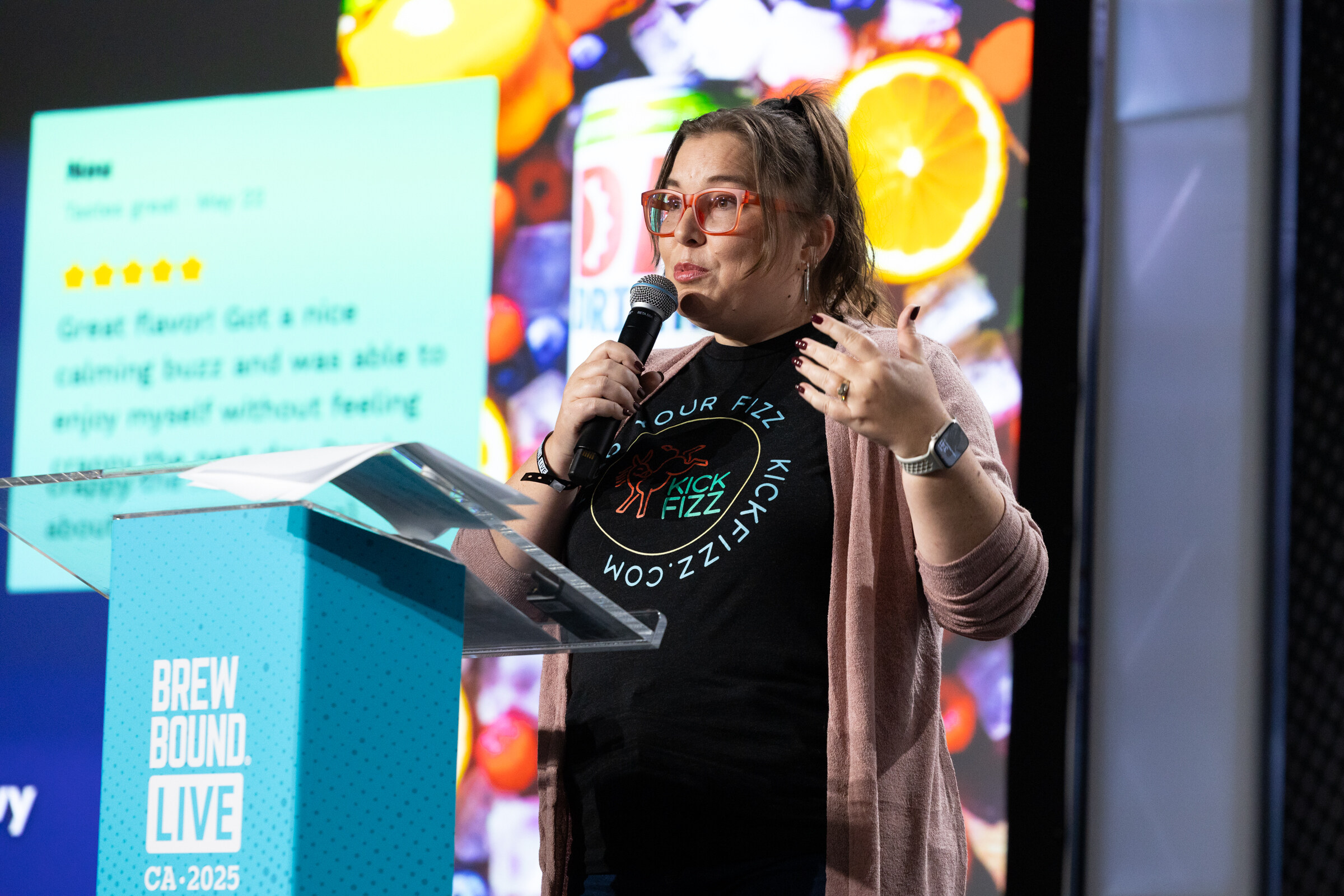 Woman in a Kick Fizz T-shirt and cardigan speaking into a microphone at a podium in front of a screen with fruit graphics.
