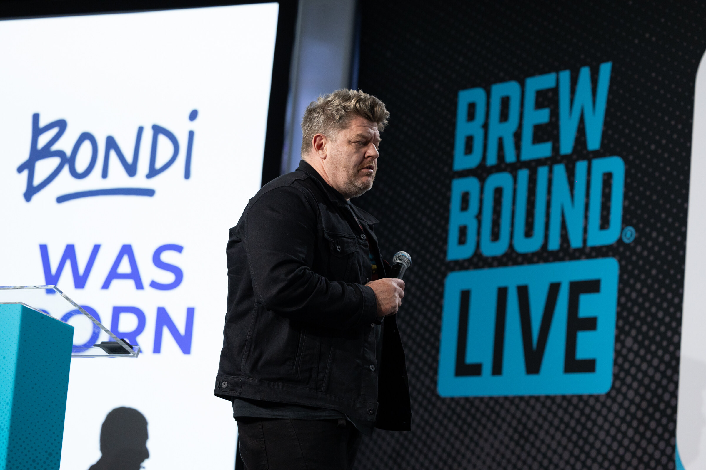 Man in a black denim jacket holding a microphone beside a podium and a white slide reading Bondi was born.