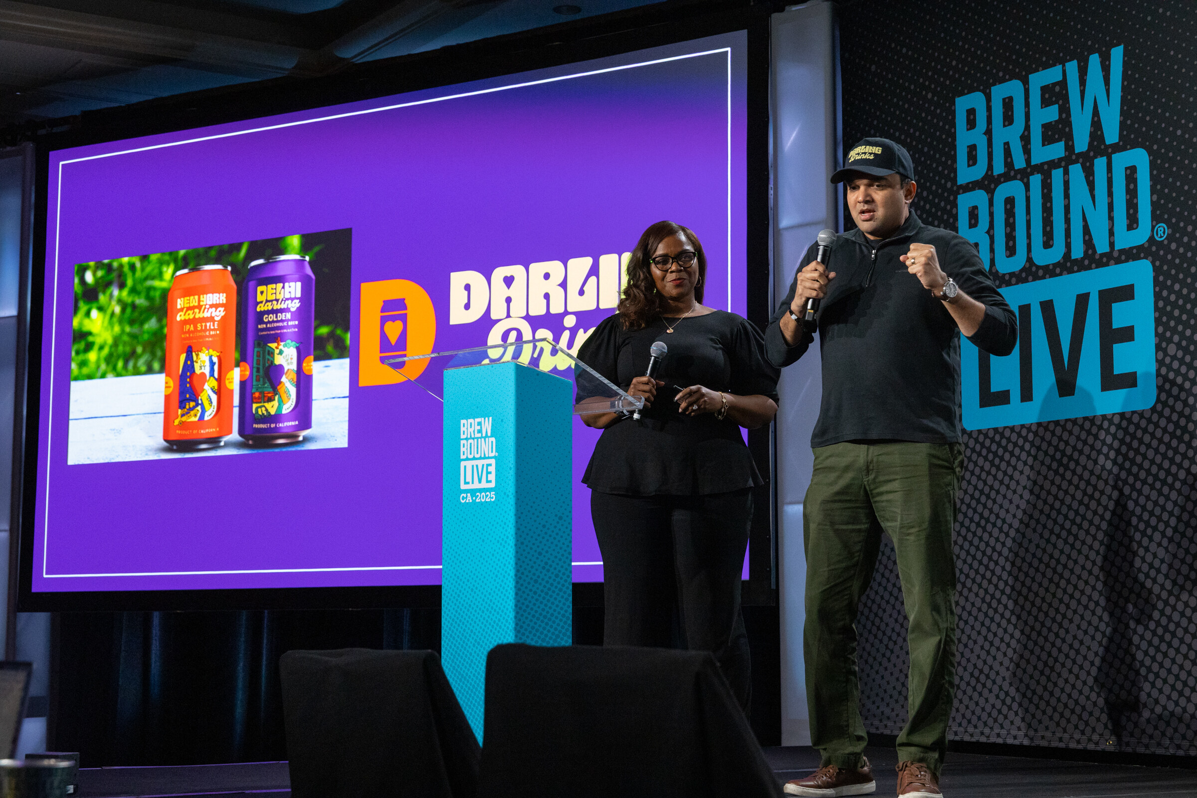 Woman in black and man in a cap and dark jacket holding microphones beside a podium and a purple screen with two cans.