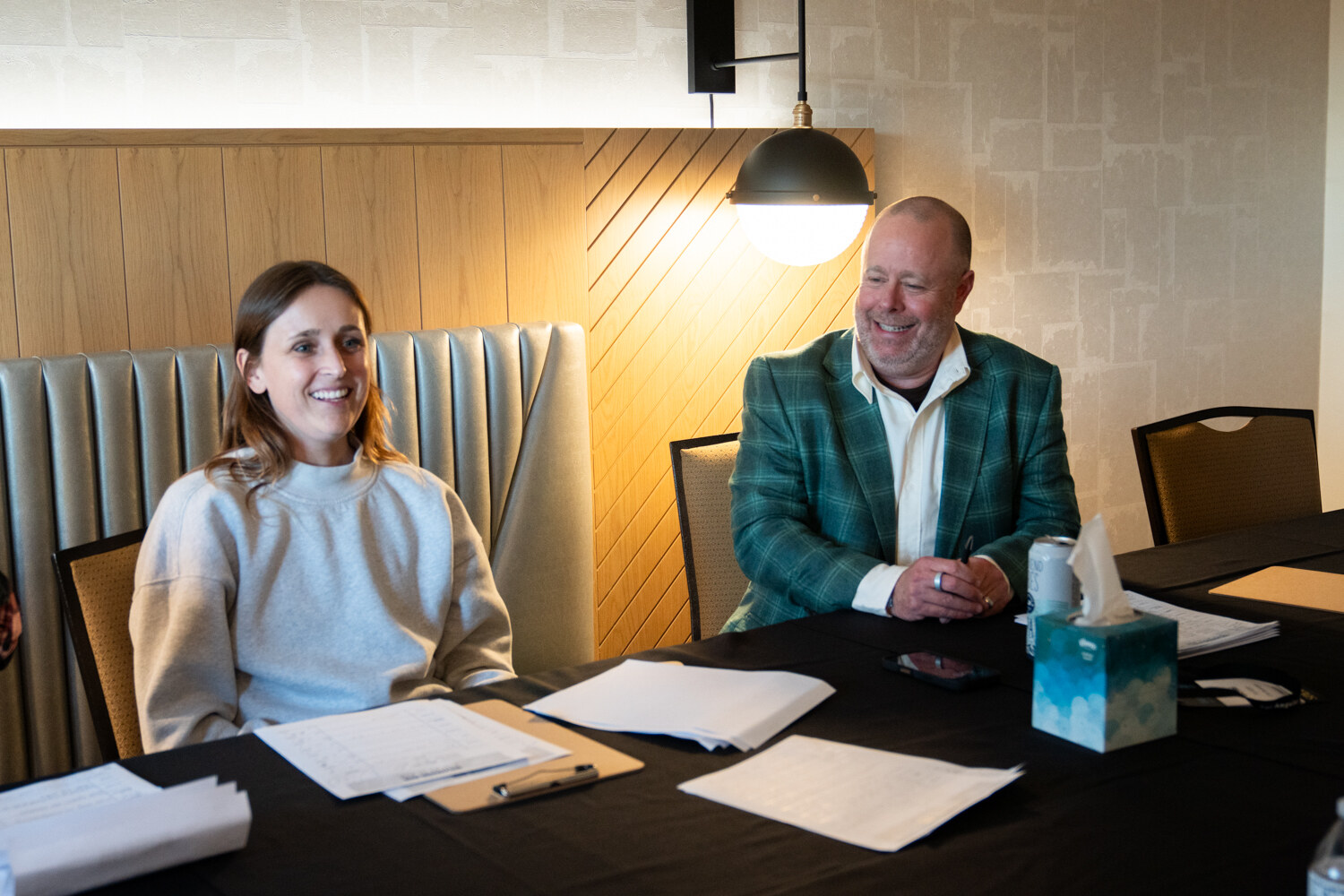 Woman in a cream sweatshirt and man in a green plaid blazer seated at a table covered with score sheets and a tissue box.
