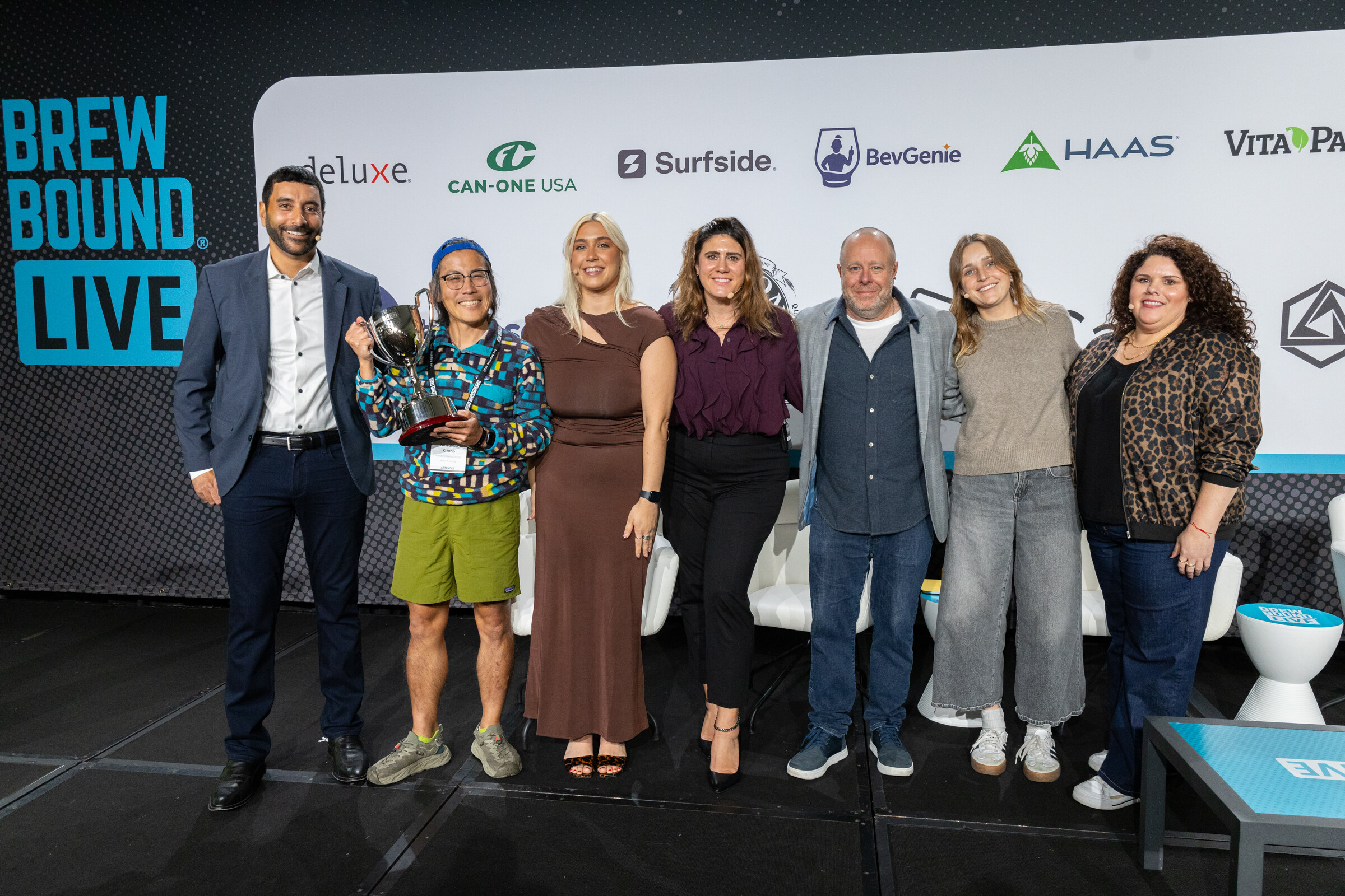 Seven people standing in a row on stage, one holding a trophy, in front of a Brewbound Live backdrop and sponsor logos.