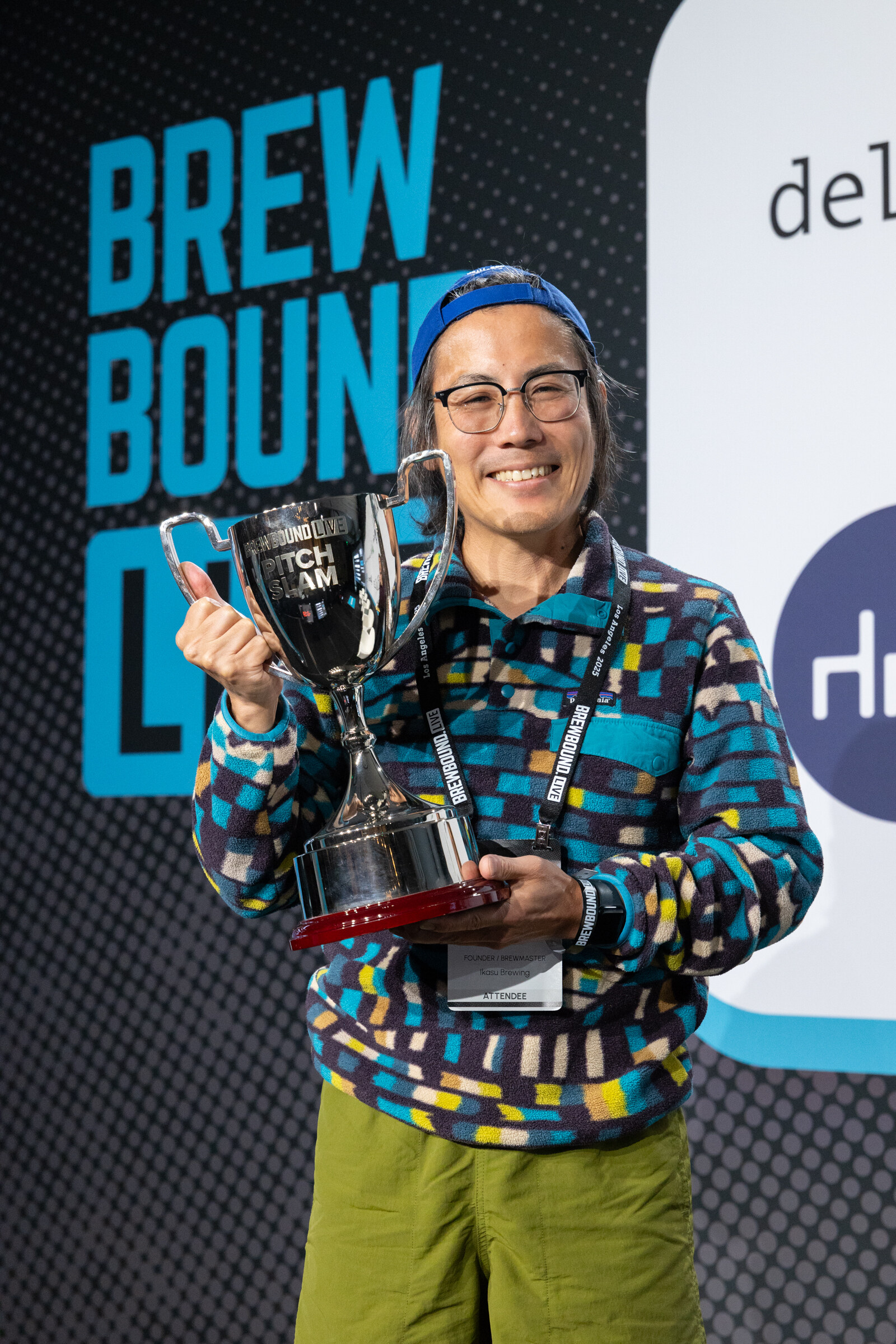Smiling man in glasses, a blue beanie and a colorful fleece holding a silver Pitch Slam trophy.